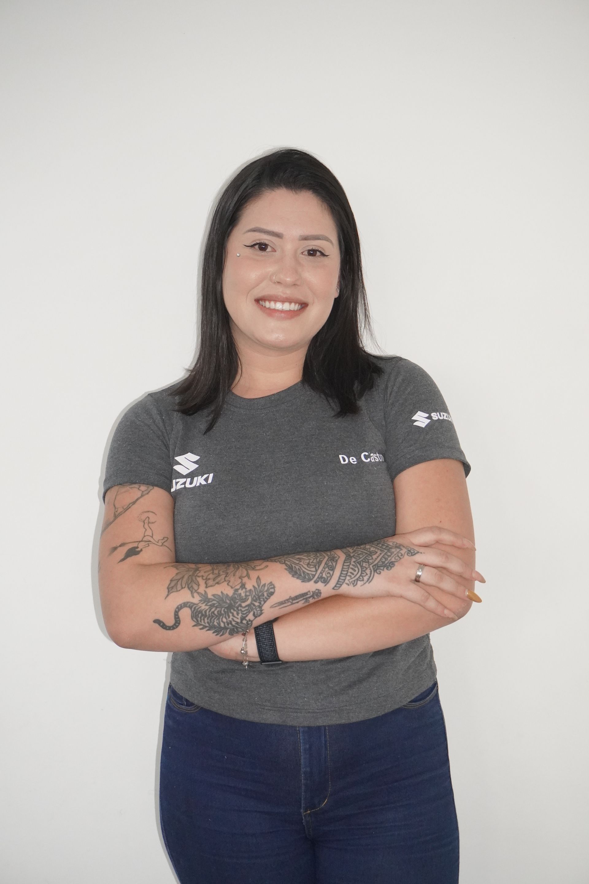 Mulher de camisa cinza e calça jeans com tatuagens no braço sorri, braços cruzados contra um fundo branco.