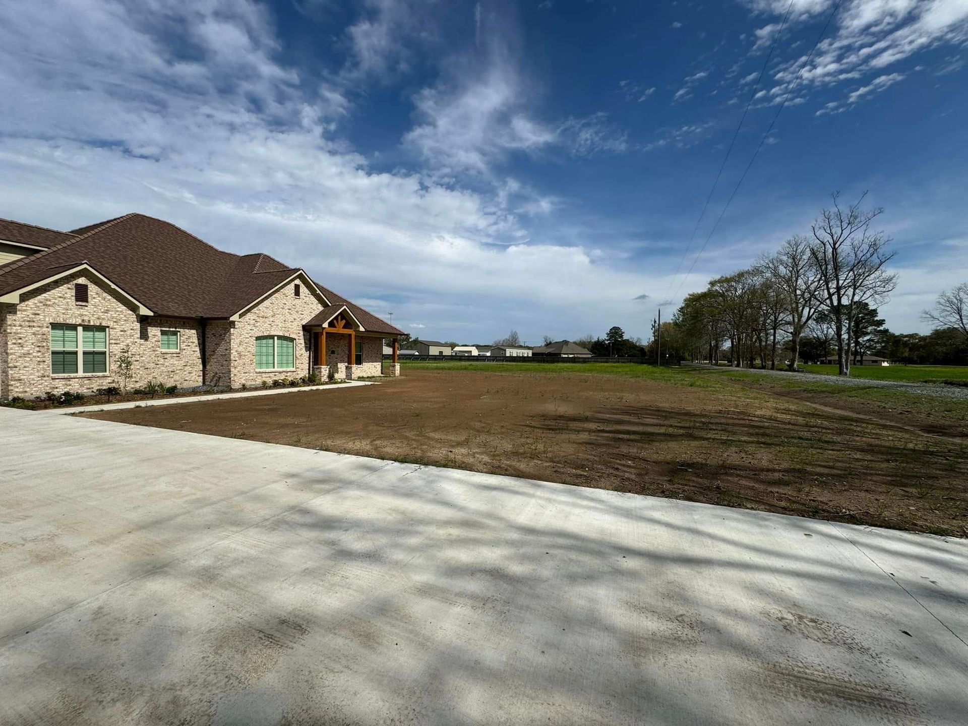 New house under construction beside a wide dirt lot and driveway on a sunny day