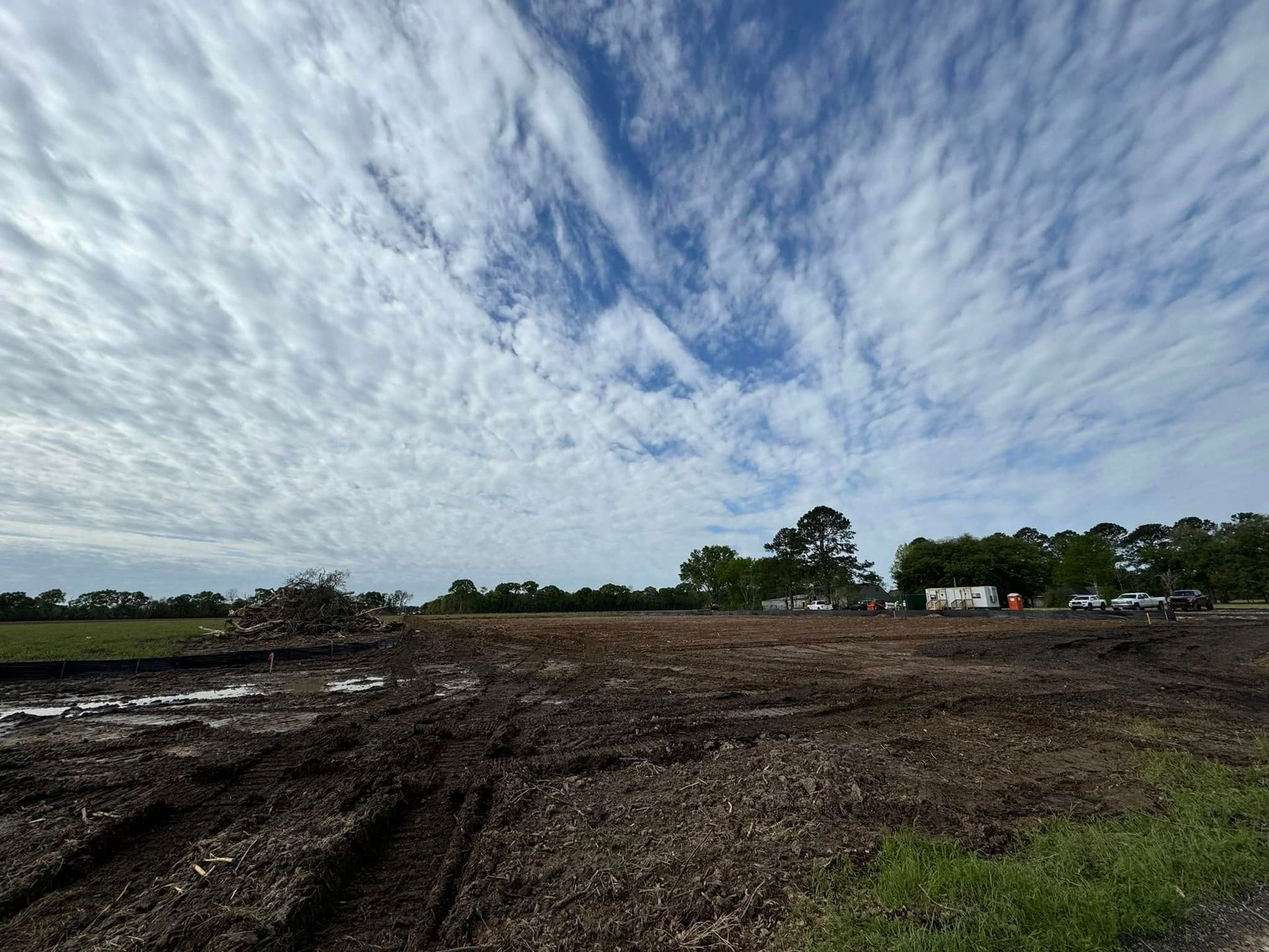 Wide muddy field under a cloudy sky with trees and a few small buildings on the horizon