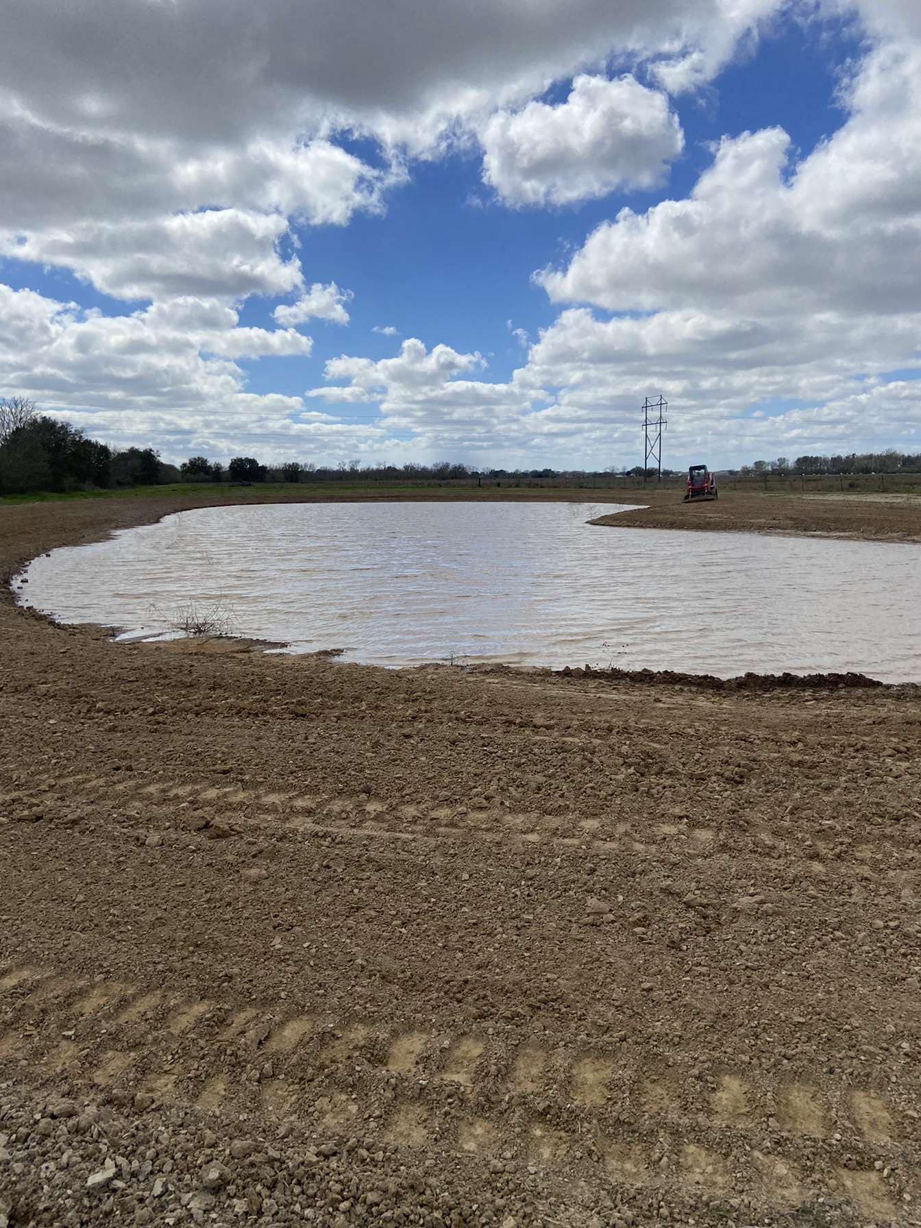 Mud-filled pond or basin under a cloudy sky, with a small pump structure in the distance.