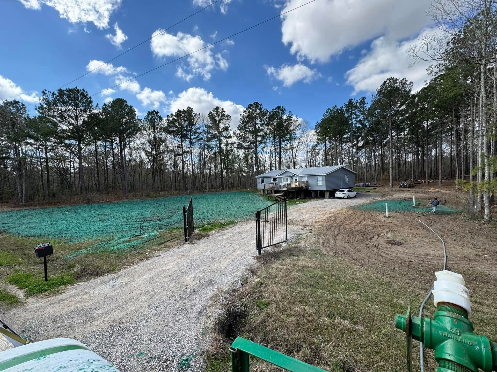 Gravel driveway leading to a gray trailer beside a green field and trees under a partly cloudy sky