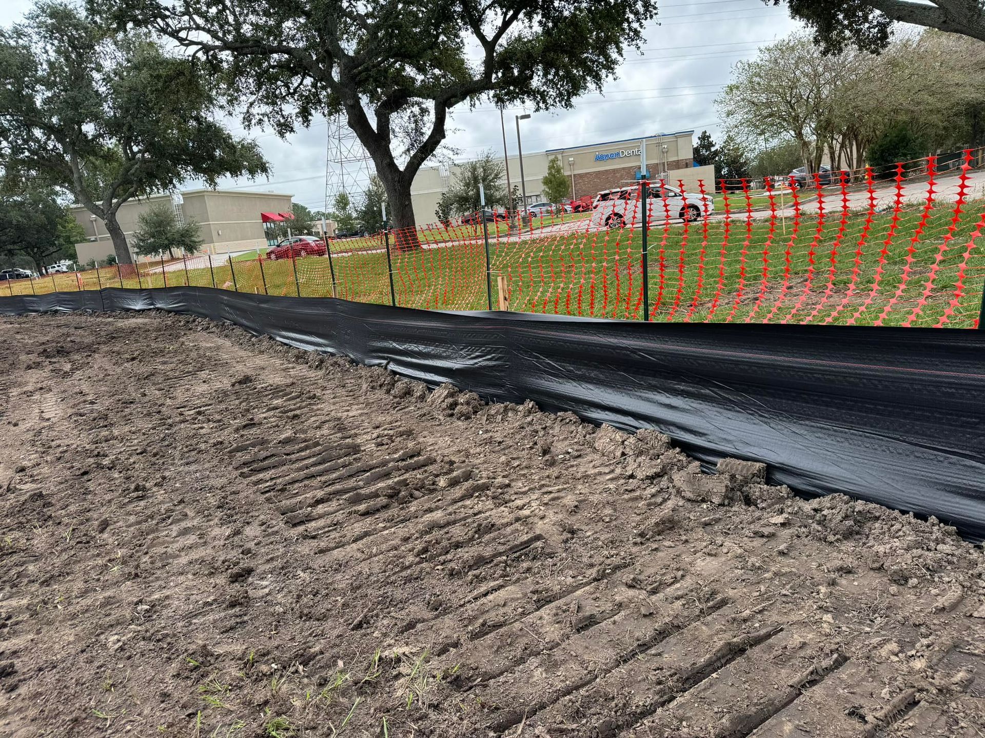 Construction site with black silt fence, dirt path, trees, and orange safety fencing in a park-like area