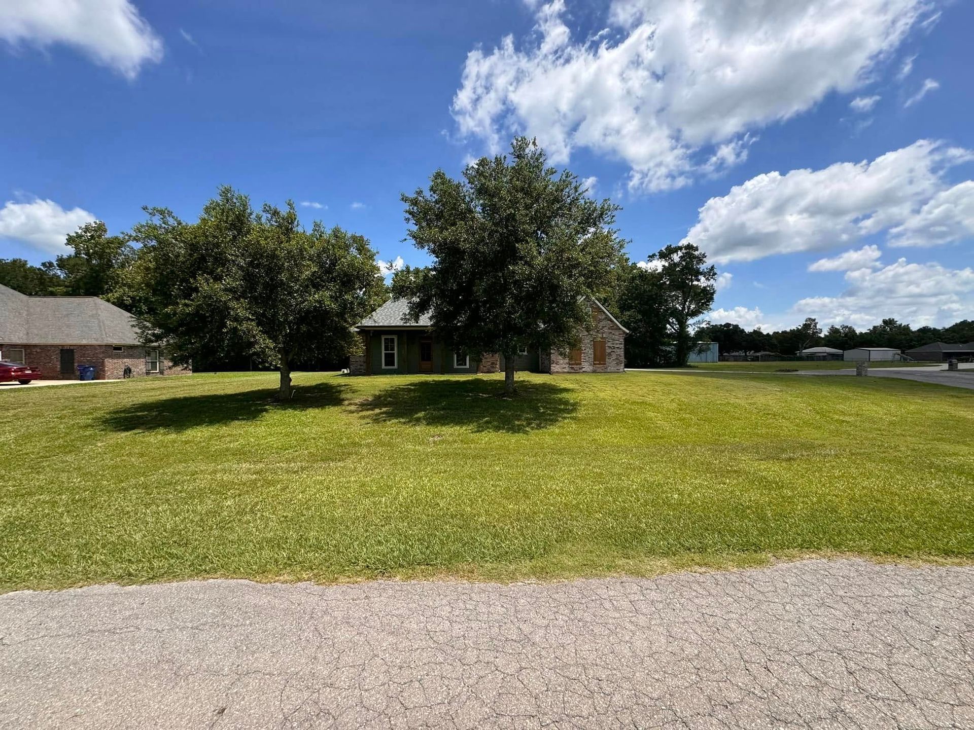 Rural yard with two trees, a house in the background, and a gravel drive under a blue sky