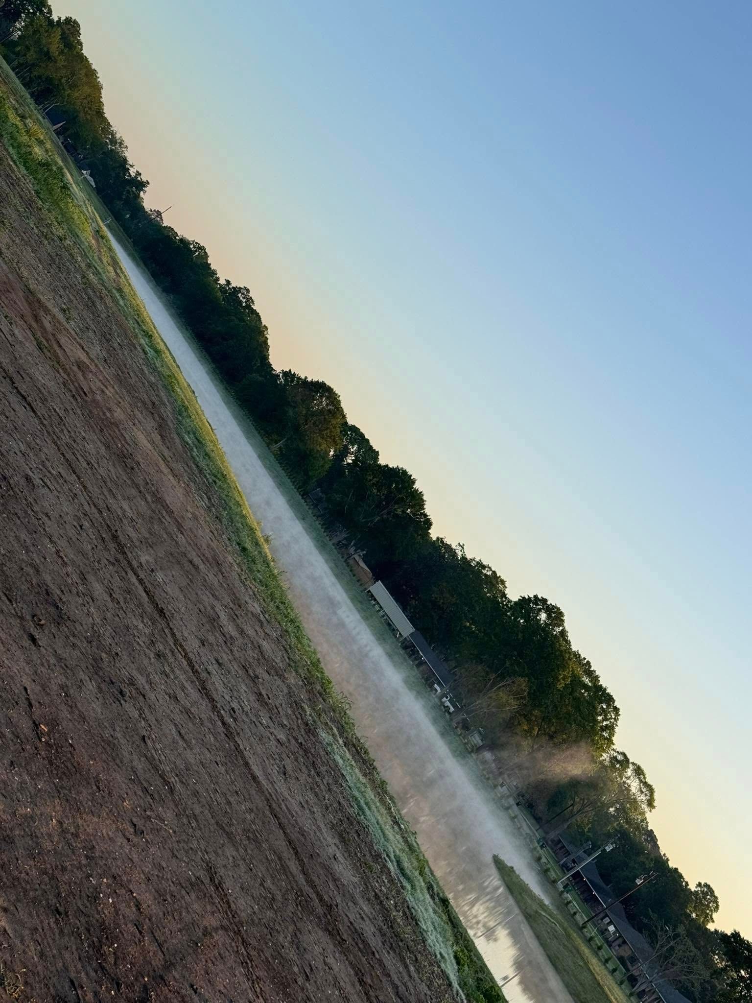 Tilted view of a dirt field with a sprinkler spraying water near a tree line at sunset