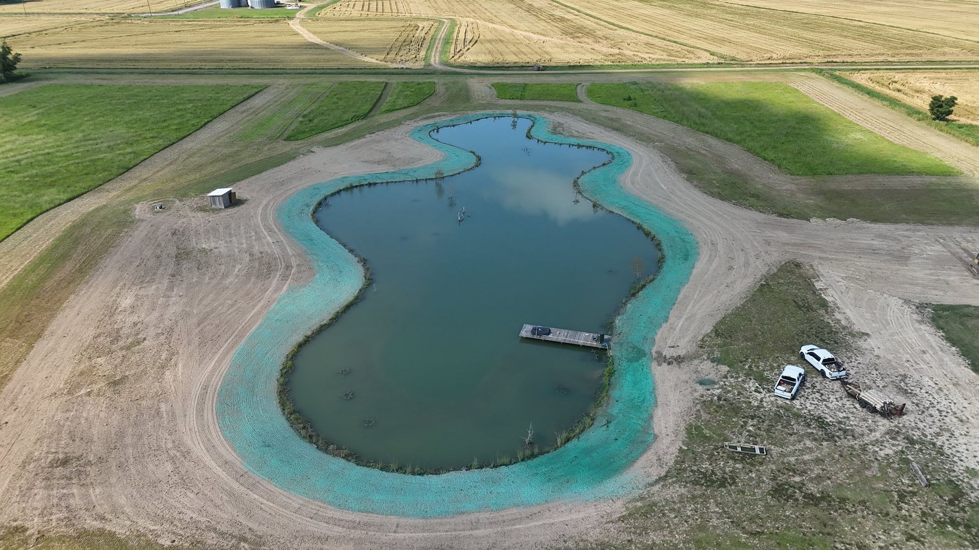 Aerial view of a small turquoise-lined pond with docks, surrounded by dry grass and fields.