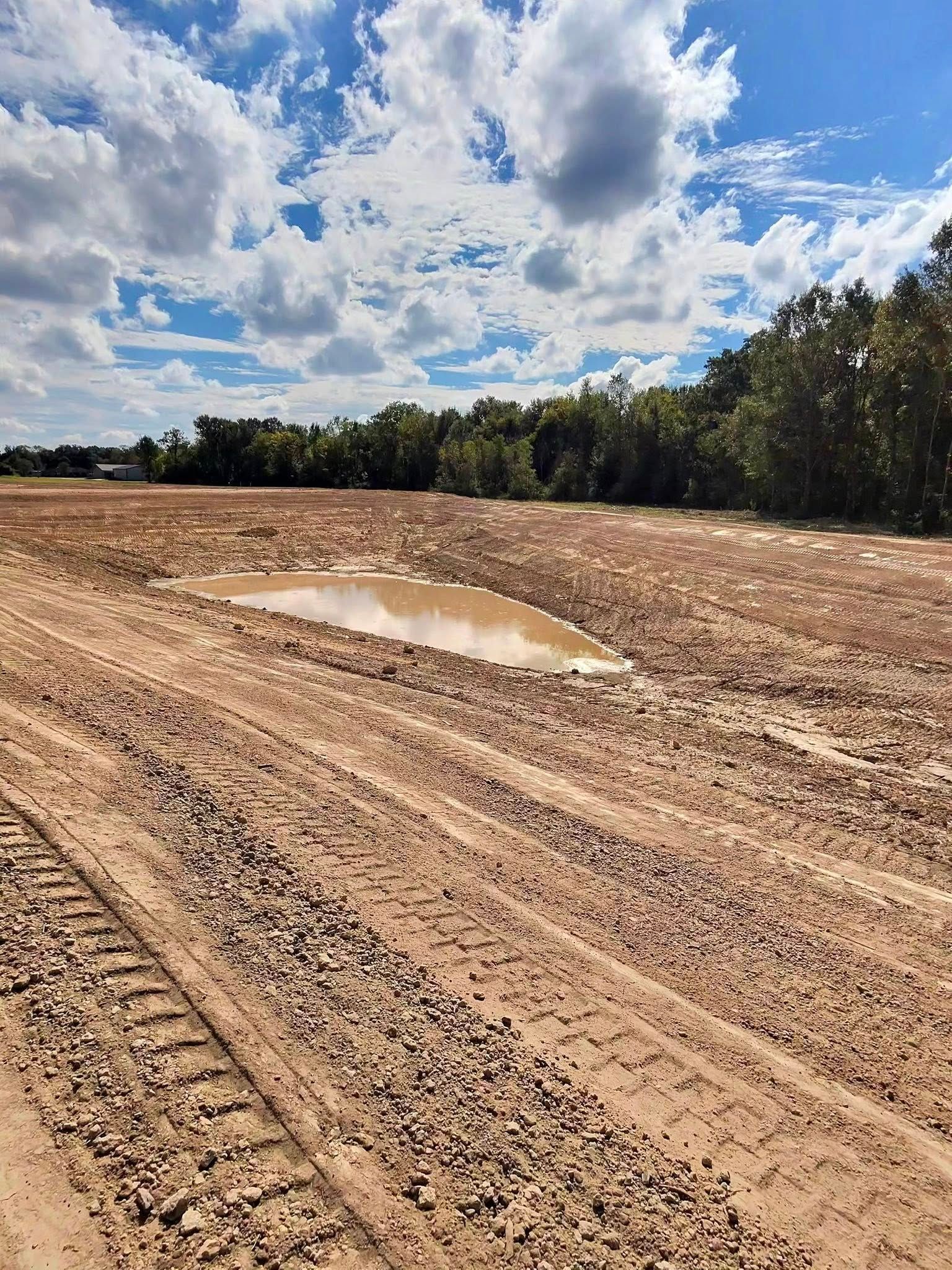 Dirt field with tire tracks and a muddy puddle under a cloudy sky, bordered by trees