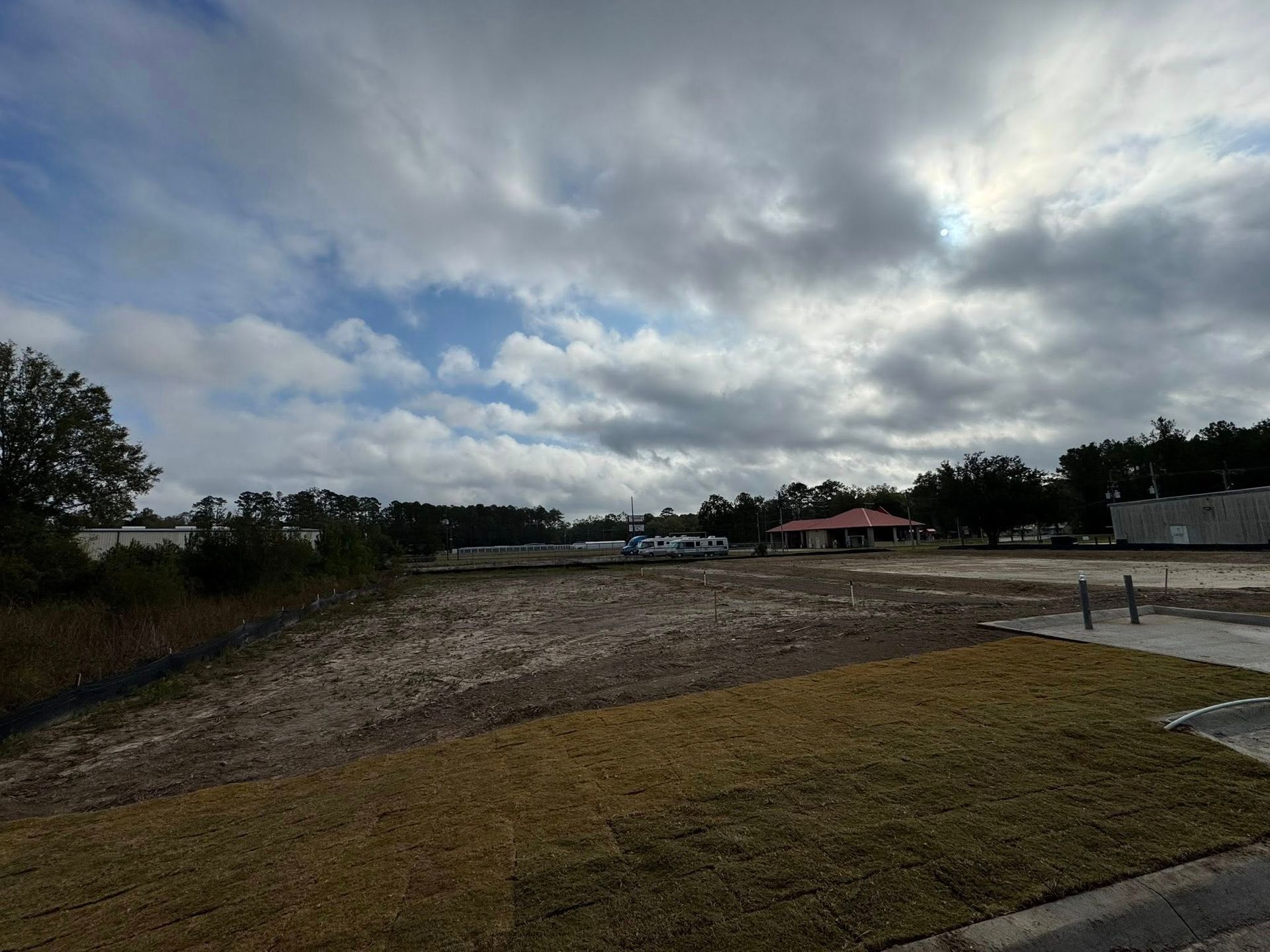Wide gravel lot beside grass under a cloudy sky, with trees and low buildings in the distance.