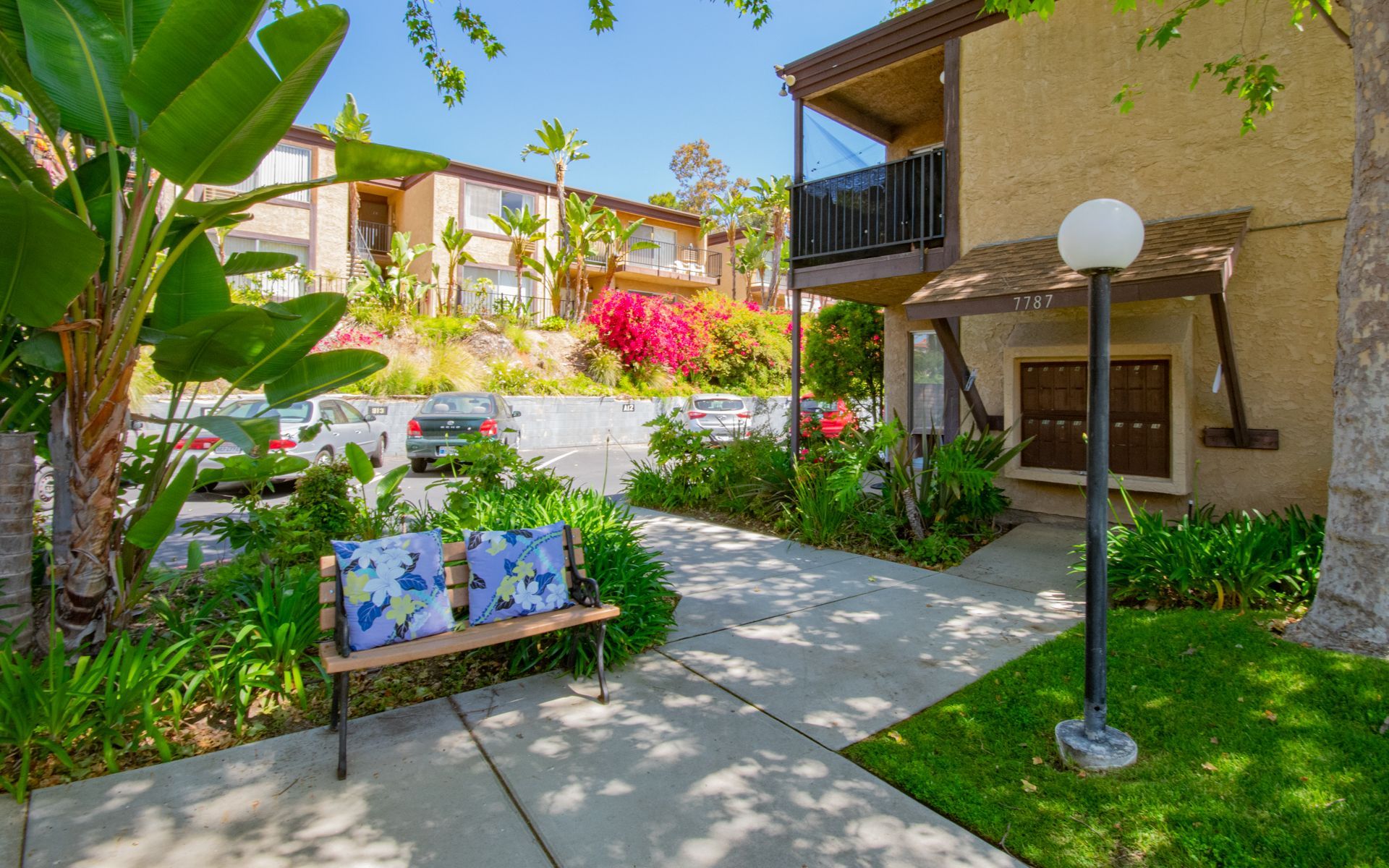 Bench on a pathway in front of a two-story building at Cowles Landing, offering apartment living in San Diego, CA. Lush greenery and other buildings visible. Sunny day.