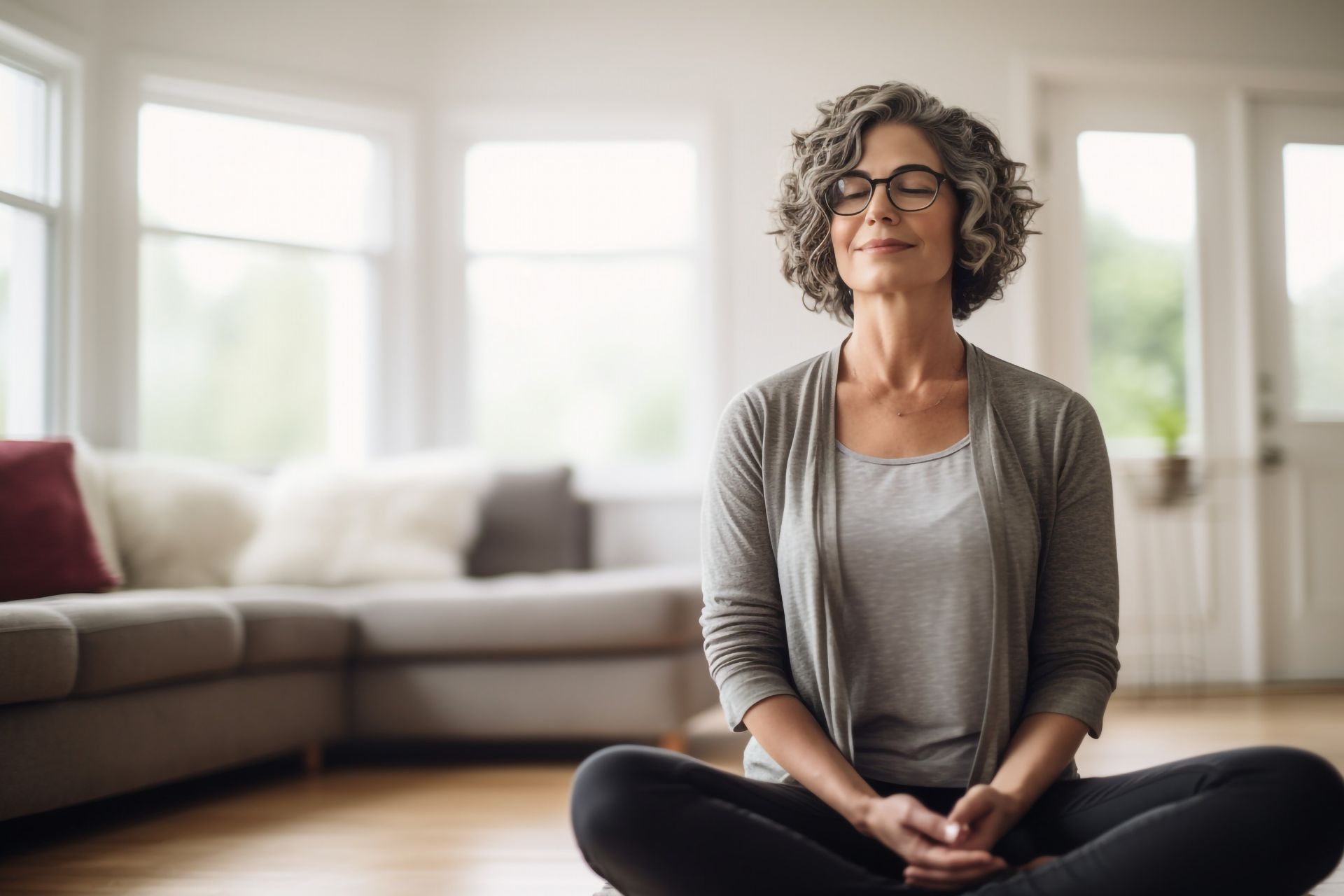 Person sitting cross-legged and meditating in a bright living room with a sofa and large windows