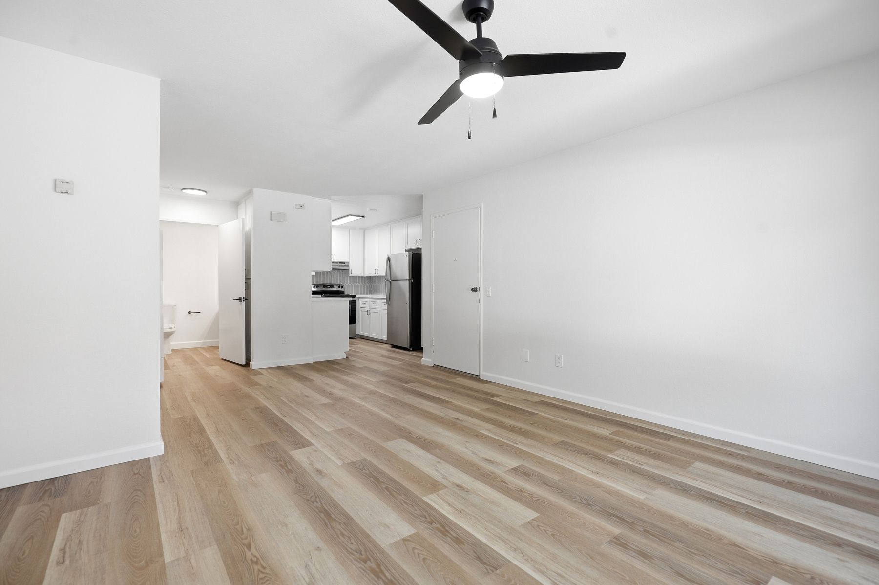 Empty modern living room with light wood floors, white walls, ceiling fan, and open kitchen area