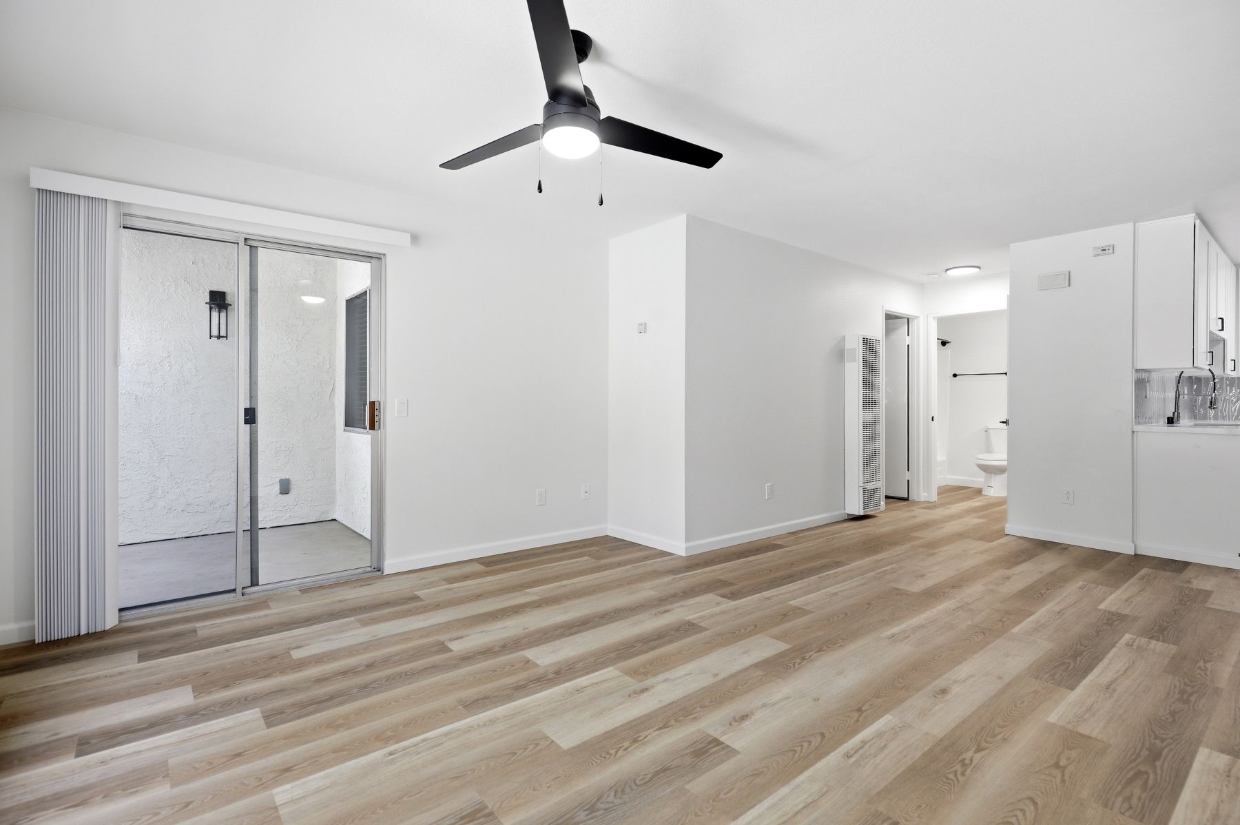 Empty modern living room with light wood floors, white walls, ceiling fan, and closet doors