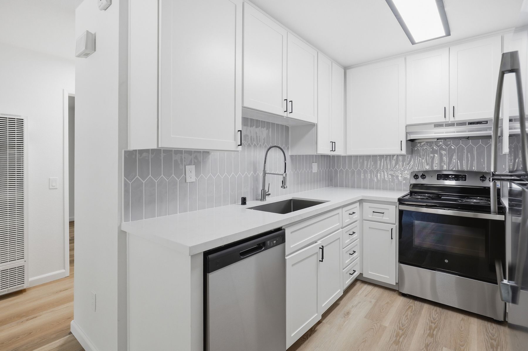 Bright white kitchen with stainless steel appliances, gray backsplash, and light wood flooring.
