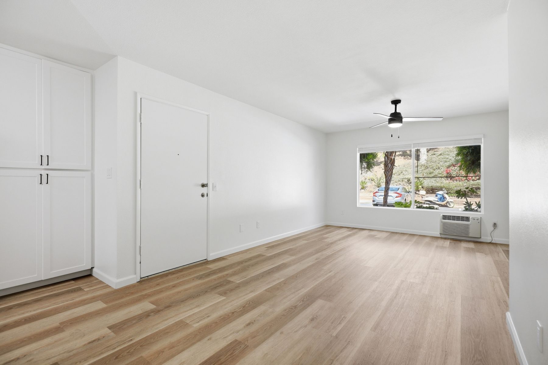 Bright empty room with light wood floors, white built-in cabinets, ceiling fan, and large window.