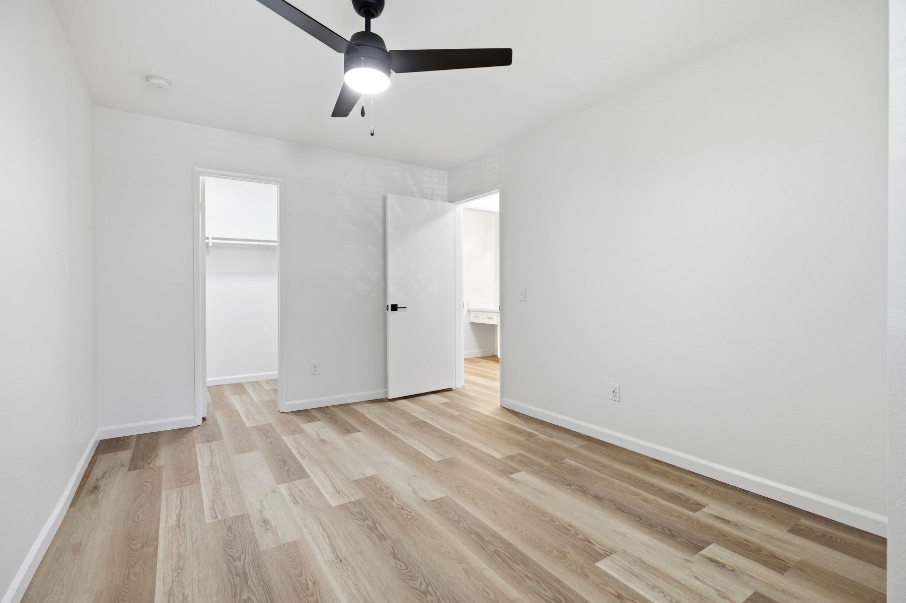 Empty white bedroom with light wood floors, ceiling fan, and open doorway to a hallway or bathroom.