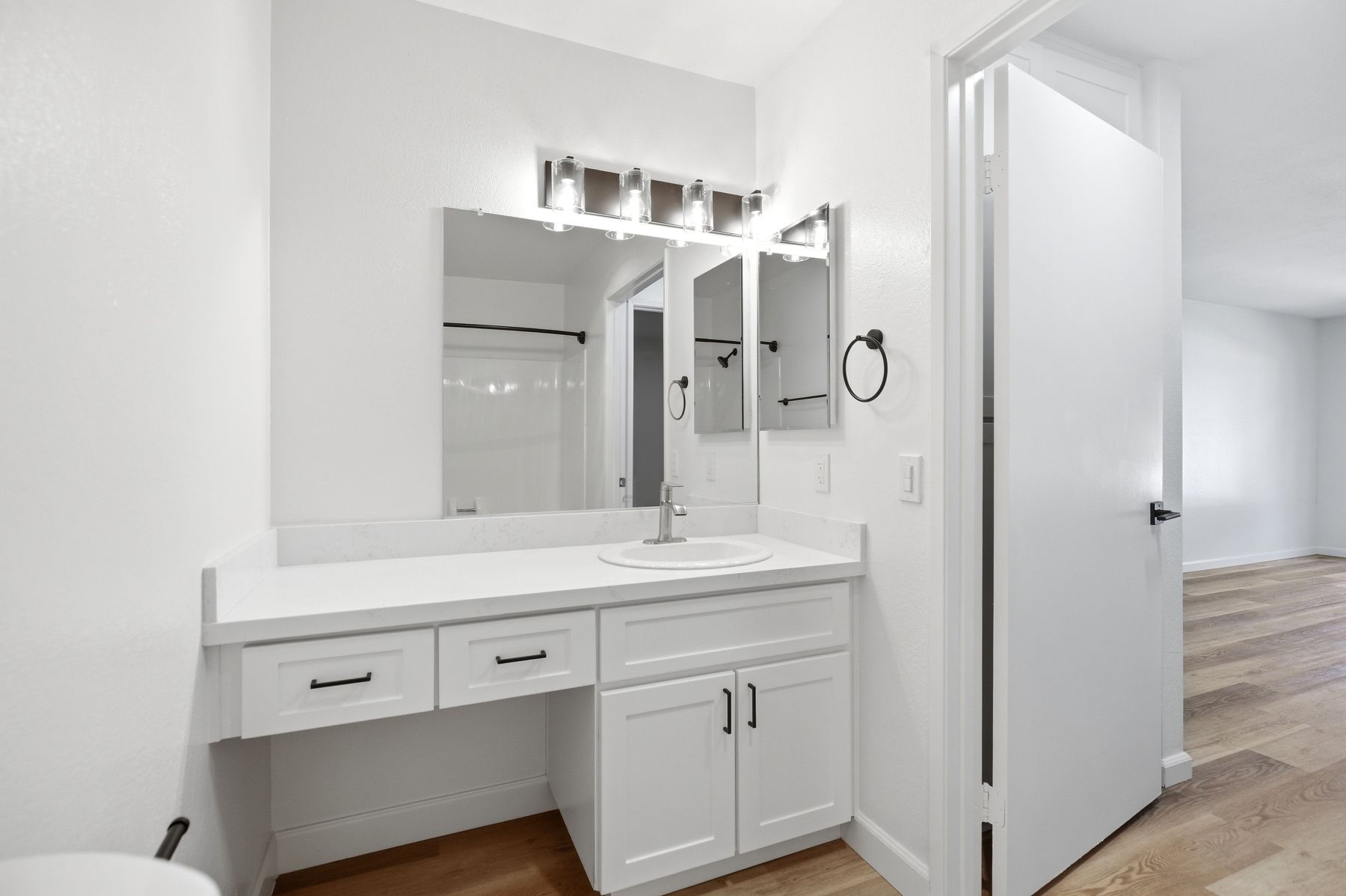 Modern white bathroom vanity with mirror, lights, sink, and open cabinet doors beside a shower and hardwood floor