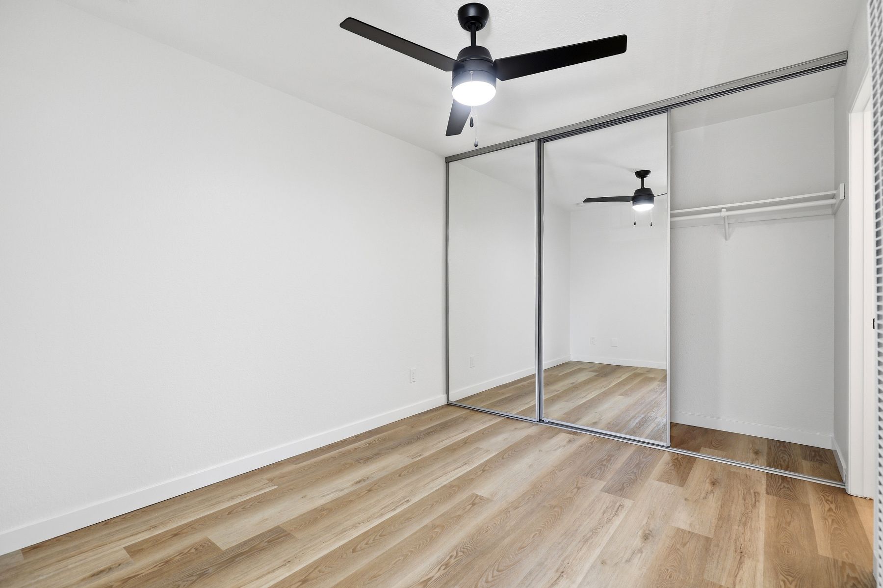 Empty modern bedroom with white walls, wood floors, mirrored closet doors, and a ceiling fan.