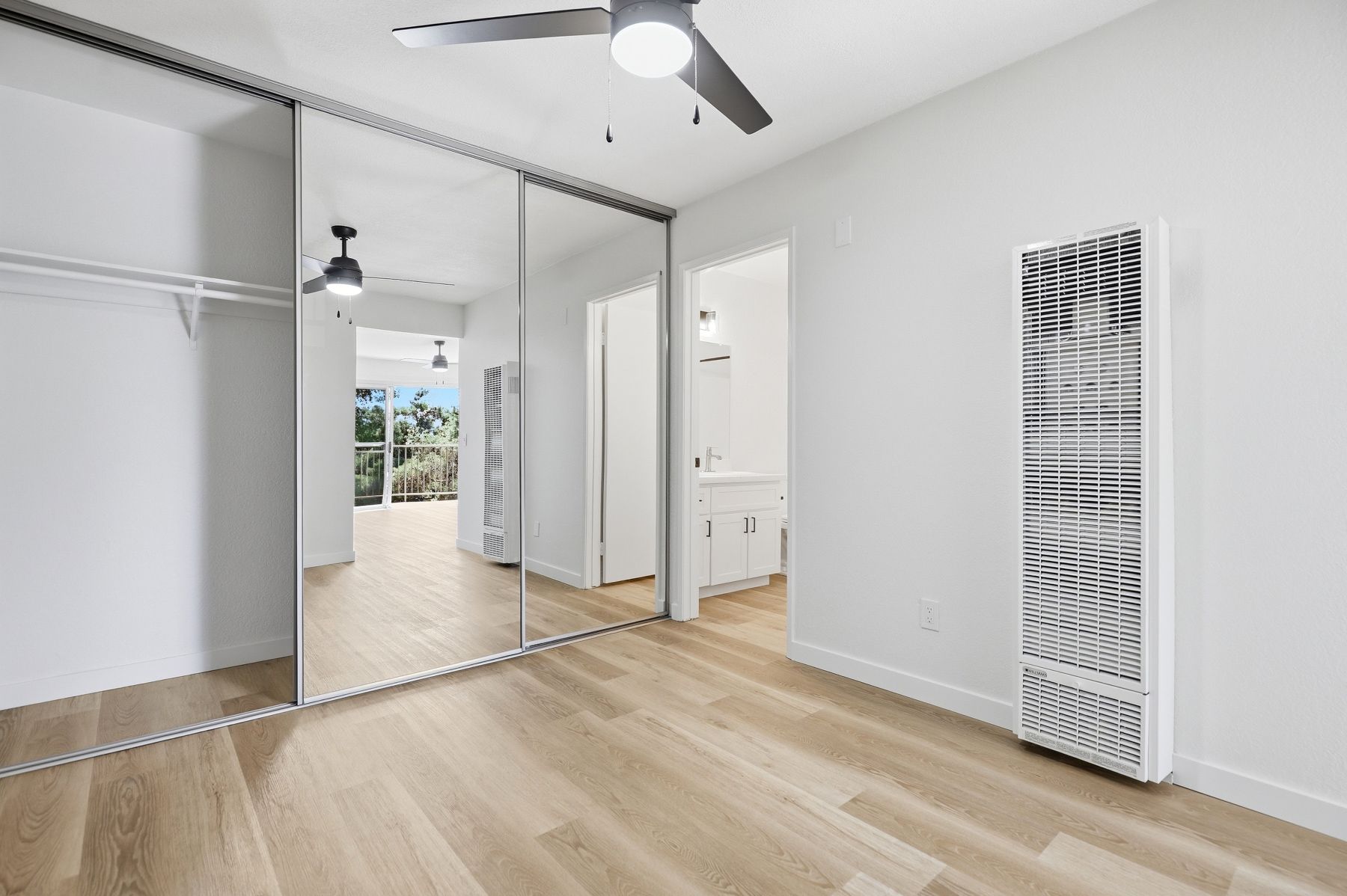 Bright empty bedroom with mirrored closet doors, light wood floors, white walls, and ceiling fans
