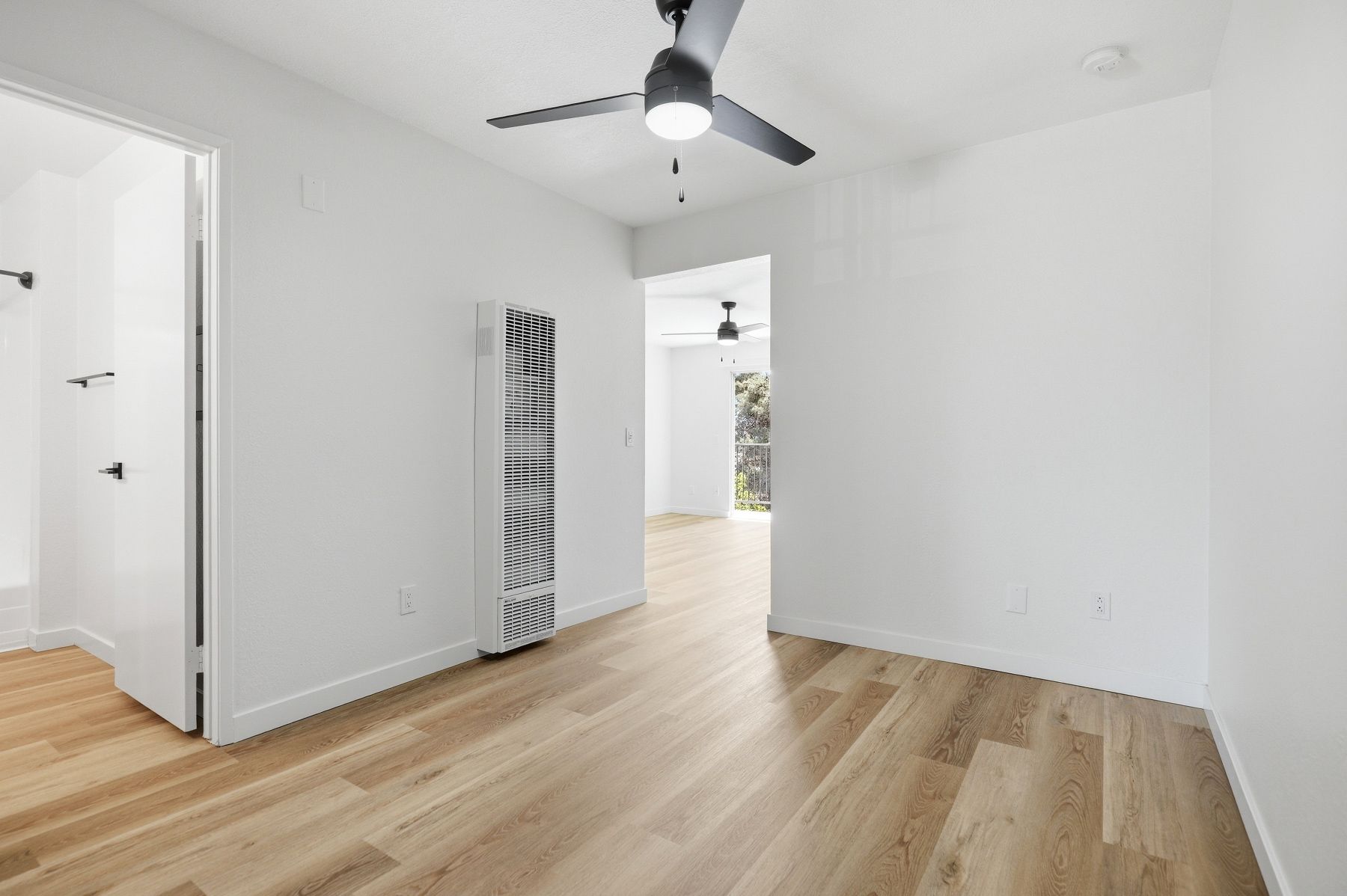 Empty white bedroom with light wood floors, ceiling fan, and open doorway to a bright hall