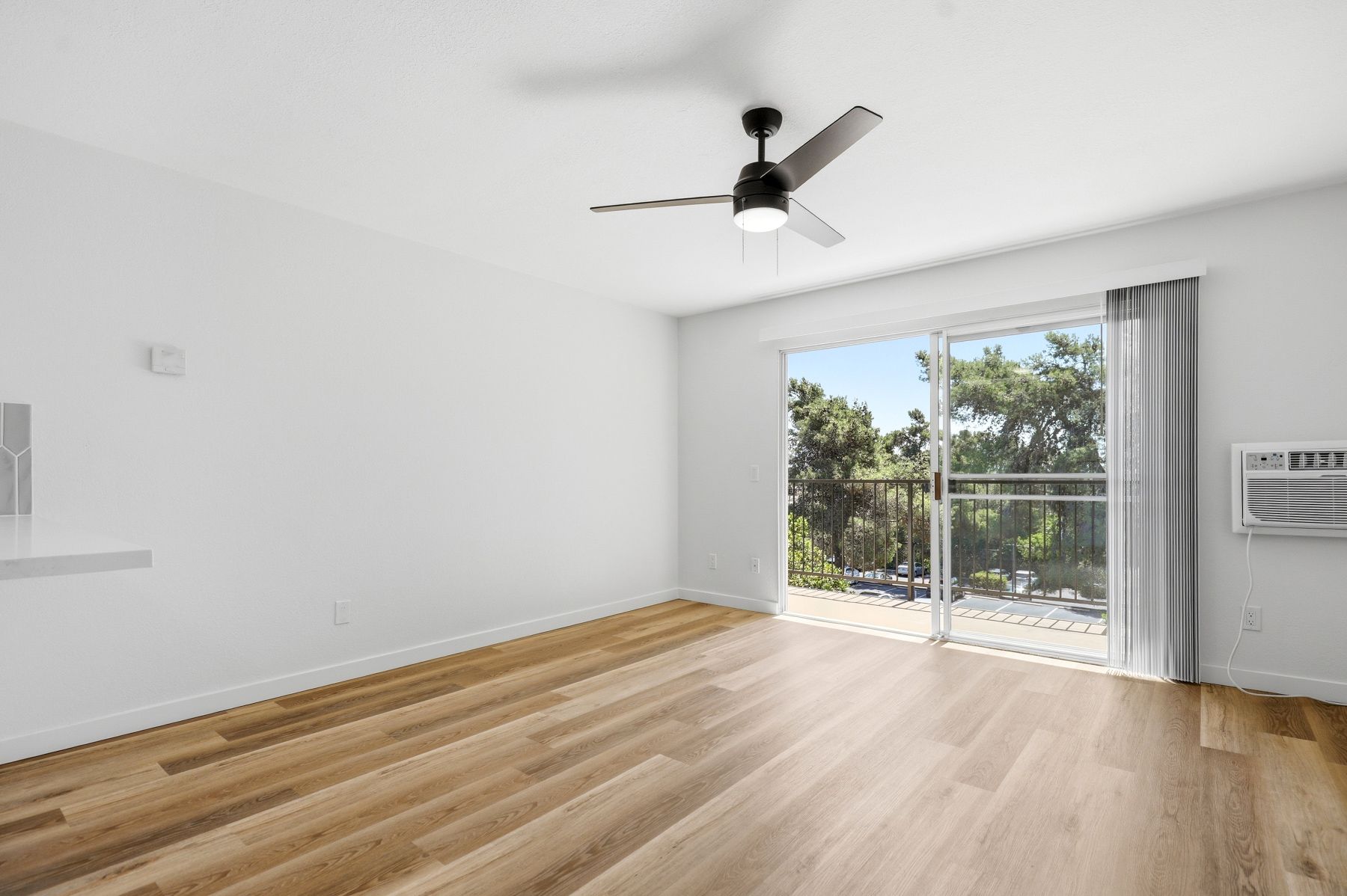 Bright empty room with wood floors, white walls, ceiling fan, and sliding glass doors to a balcony.