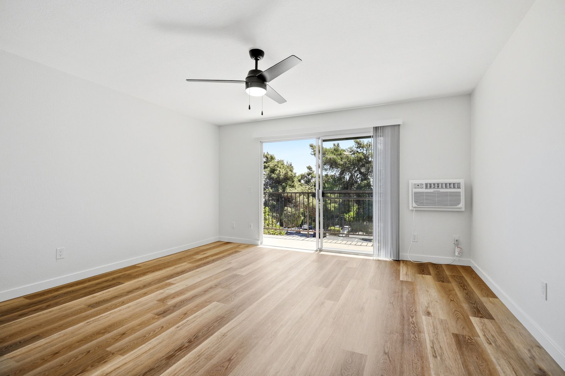 Bright empty room with wood floors, ceiling fan, and sliding glass door to greenery