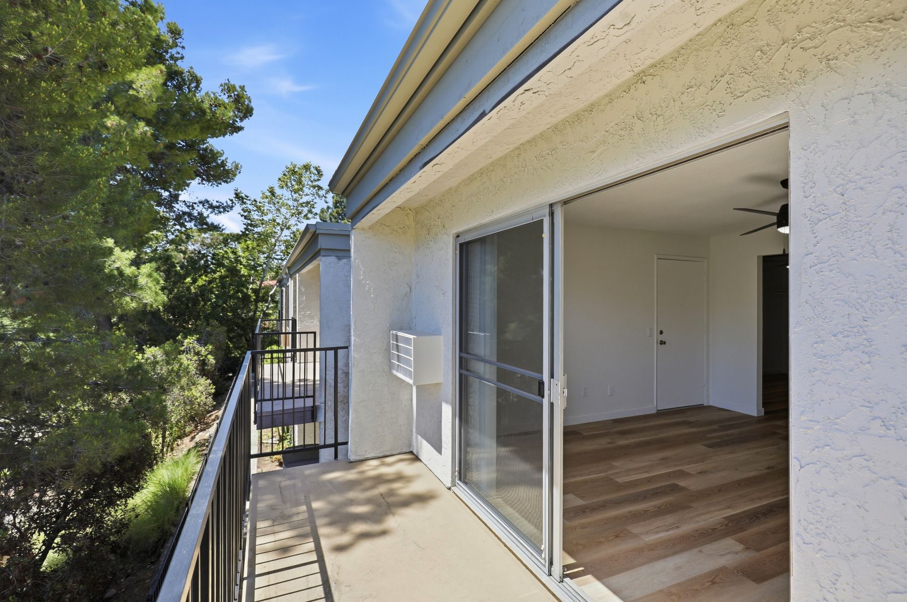Balcony beside an open sliding glass door, with trees and a bright wood-floored interior visible.
