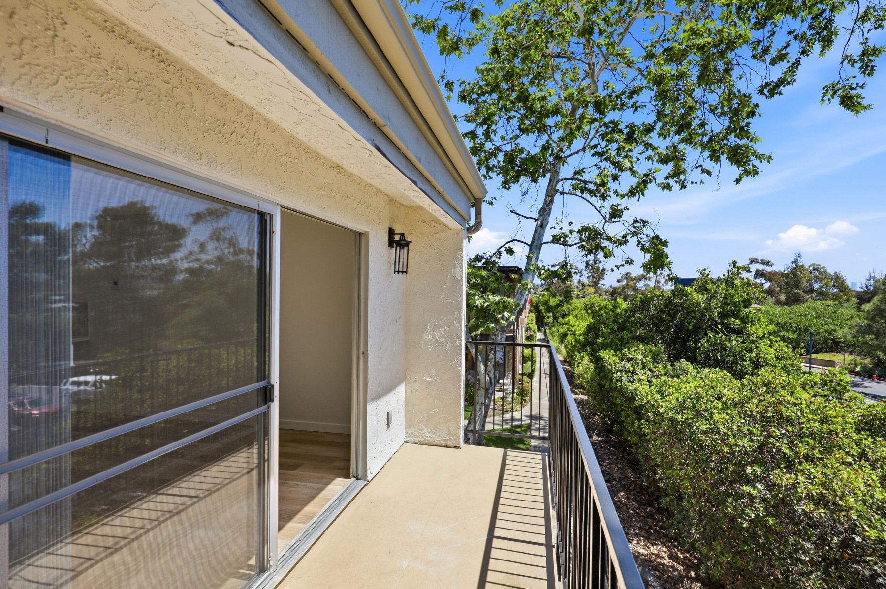 Balcony walkway beside a beige building with glass door, railing, and green trees beyond