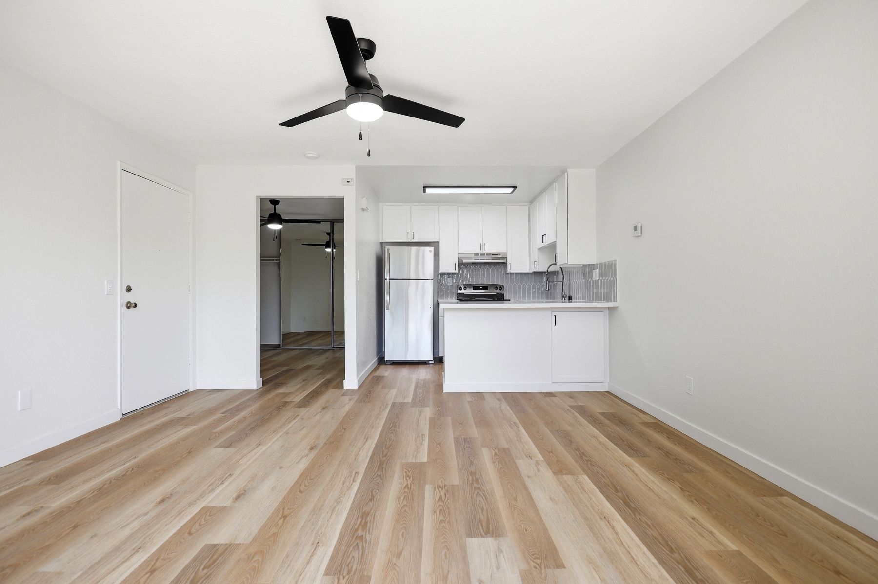 Empty modern apartment with light wood floors, white walls, ceiling fan, and small kitchen area