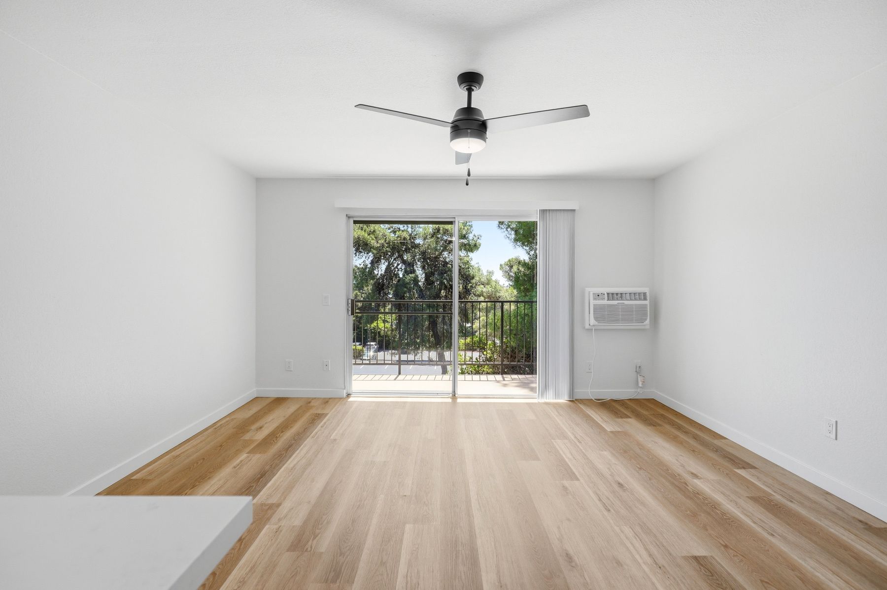 Empty white living room with wood floors, ceiling fan, and sliding glass doors to a patio.