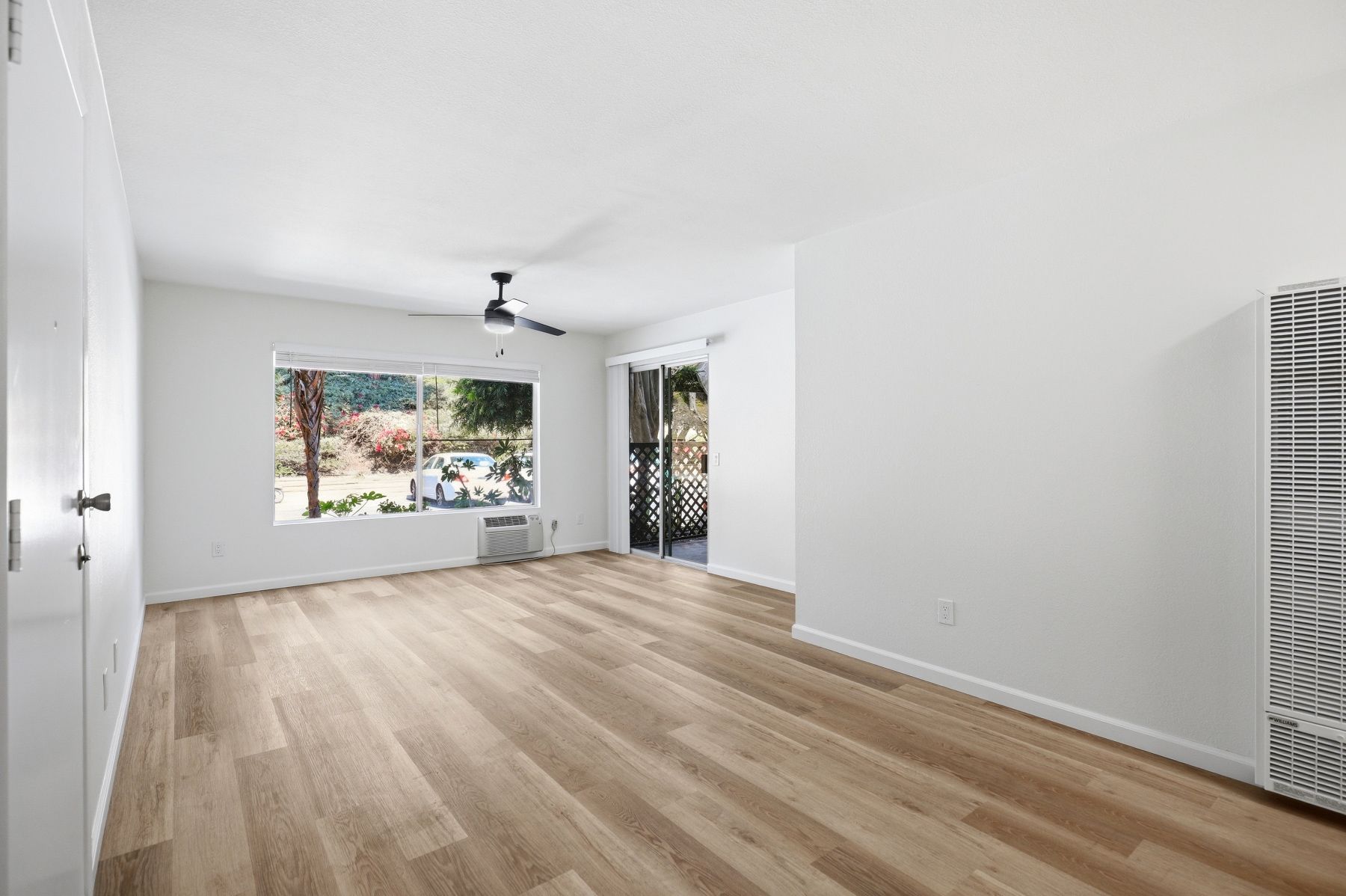 Empty modern living room with white walls, light wood floors, and large windows overlooking greenery