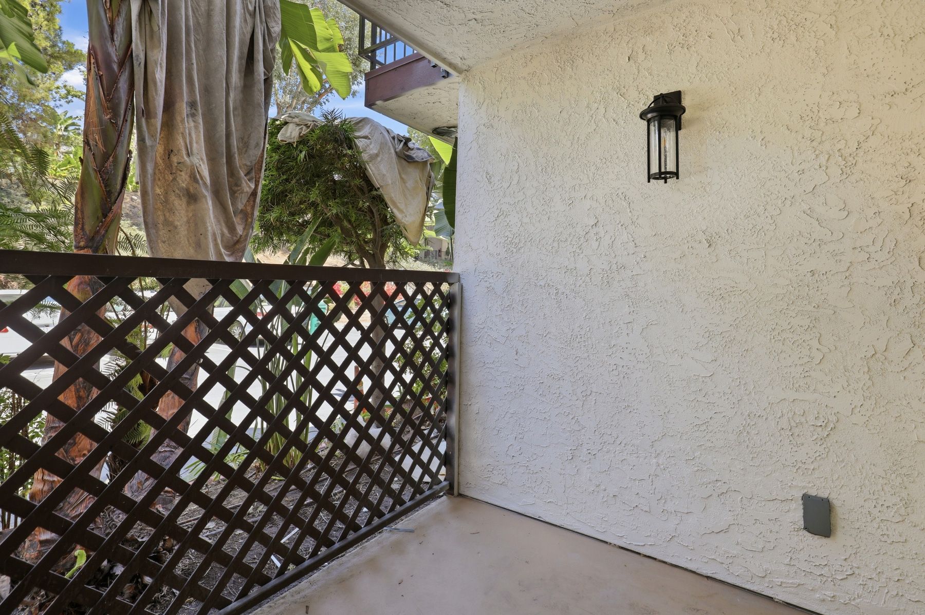 Small patio with white stucco wall, lantern light, and dark lattice fence beside palm trees.