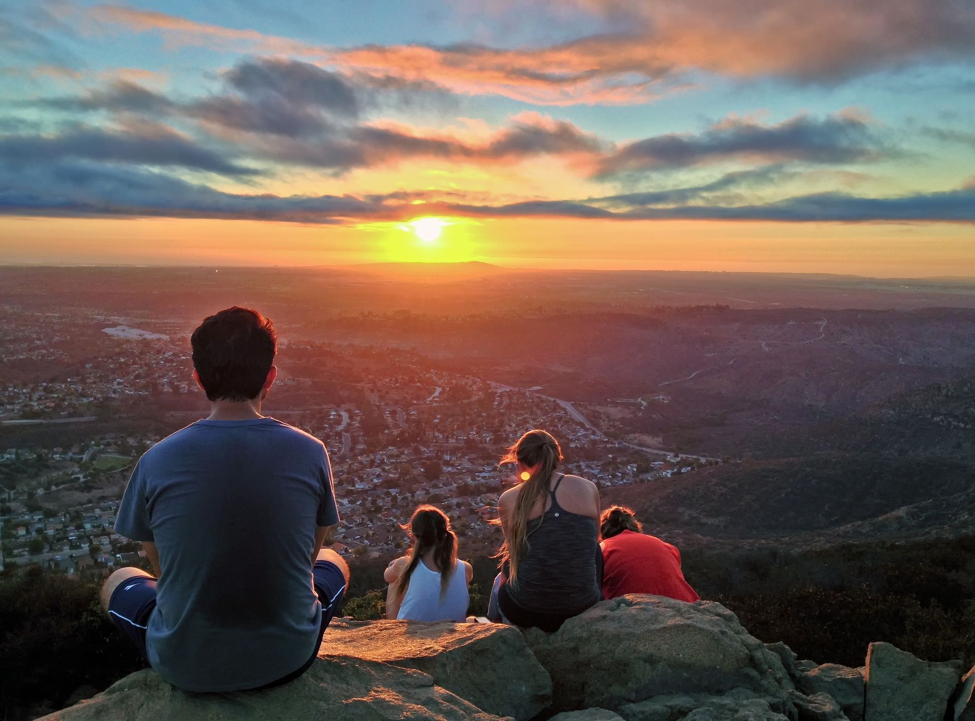 Four people sit on a rocky overlook at sunset, watching the glowing sky over a wide valley.