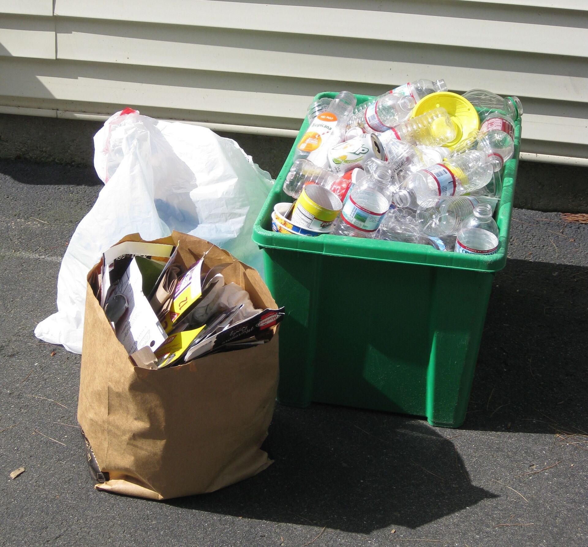 Contenedores de reciclaje con botellas de plástico transparente, bolsas de papel y una bolsa blanca en un camino de entrada.