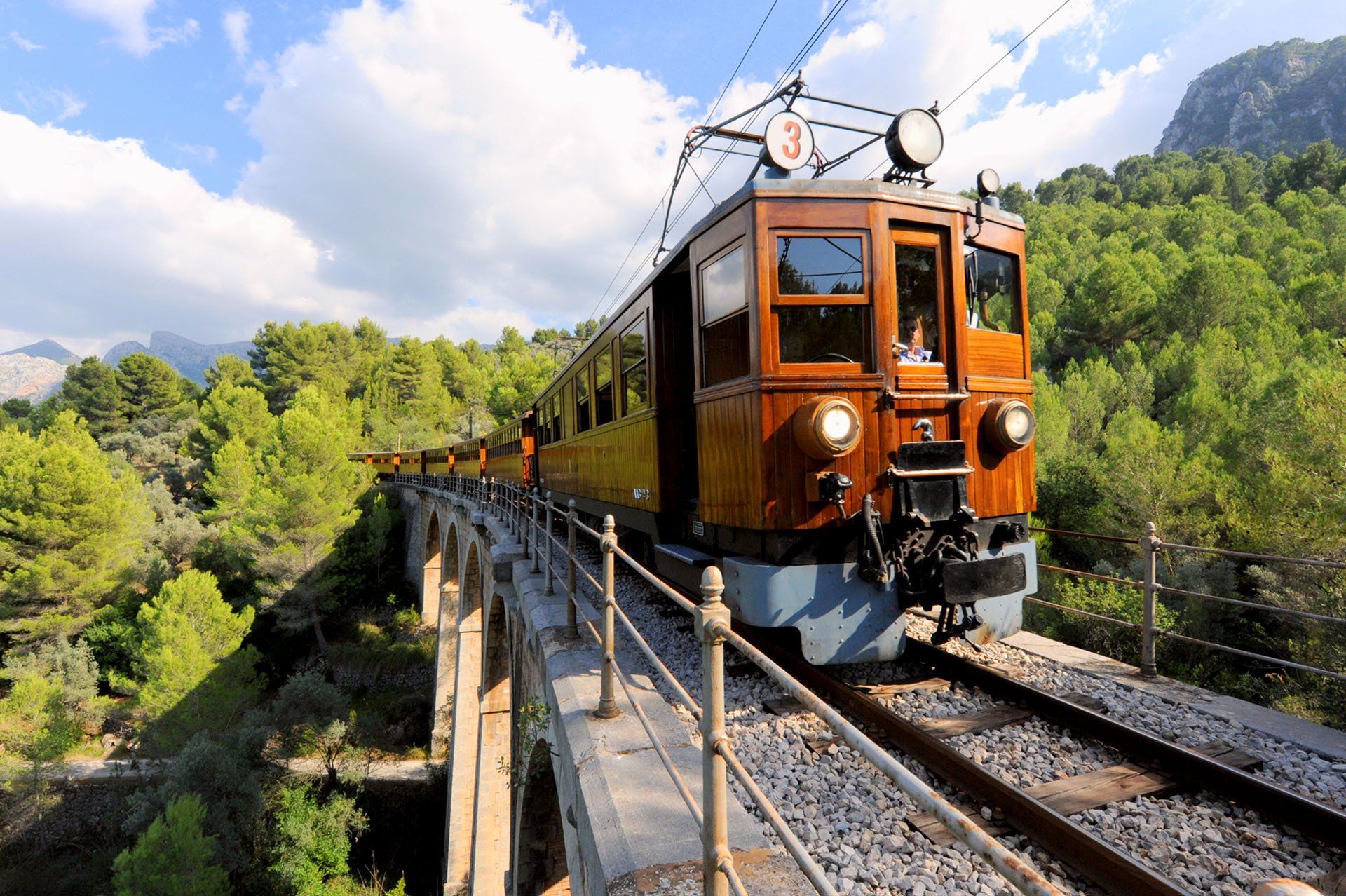 Tren antiguo de madera viajando sobre un puente, con montañas y árboles al fondo.