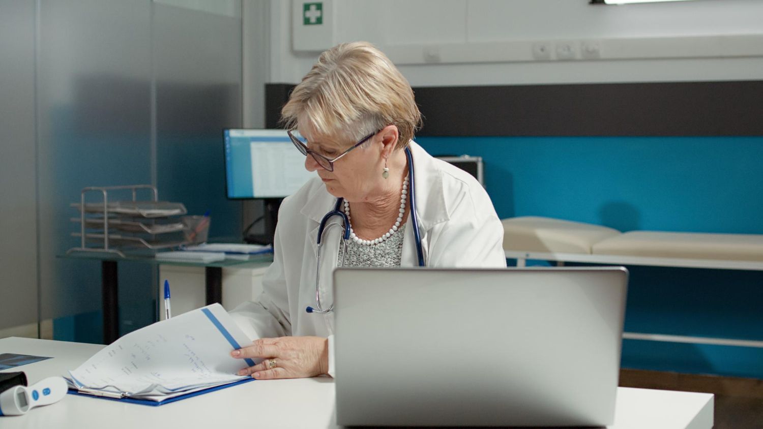 Doctor writing notes at a desk in a clinic, with a laptop and open folder in front of her.