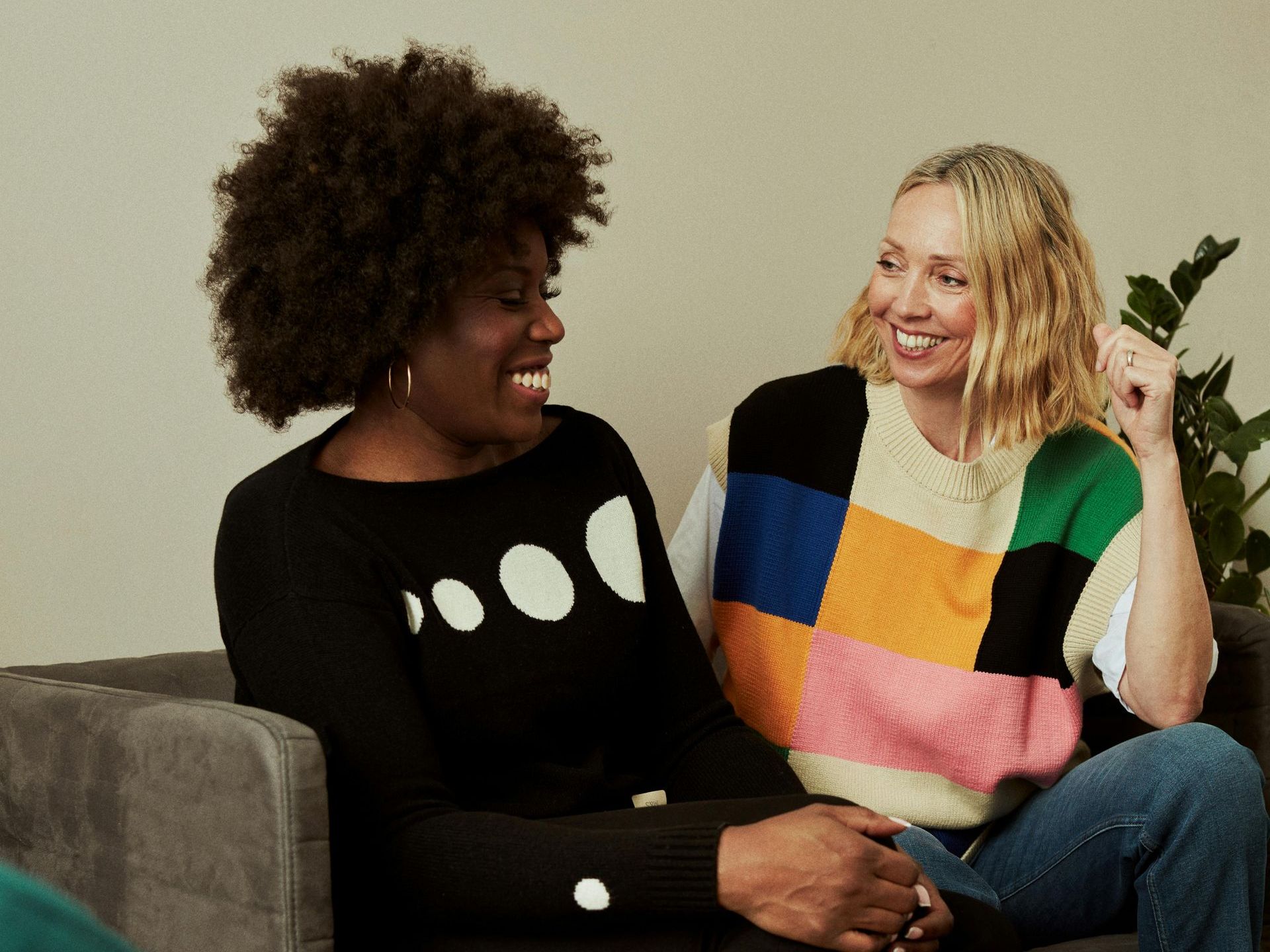 Two people smiling on a couch, one in a black top and one in a colorful sweater.