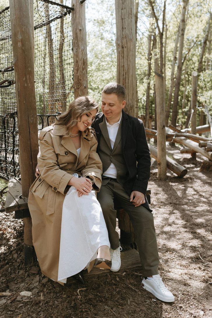 Two people sitting close together on a bench in a wooded outdoor setting, smiling at each other.