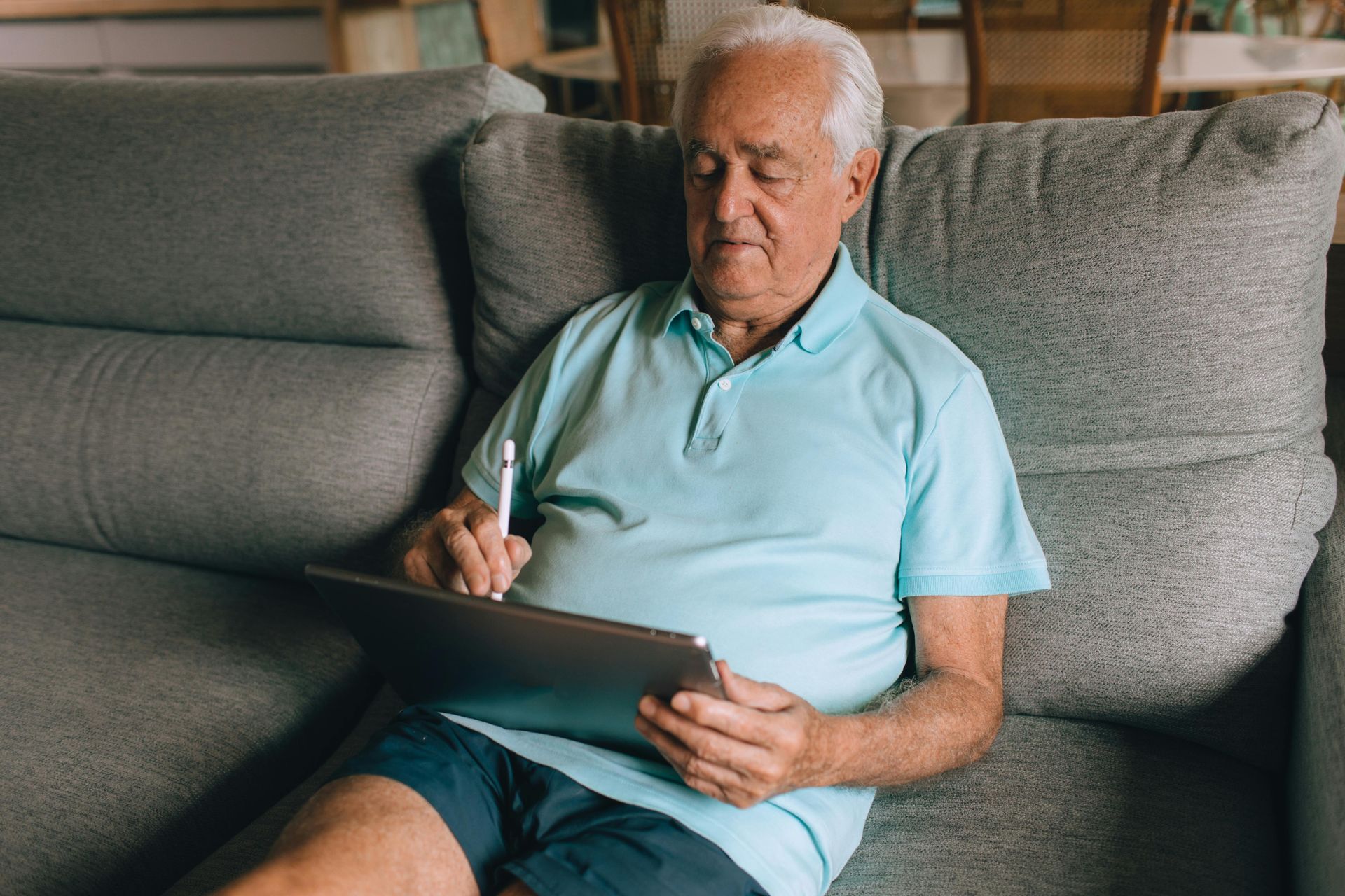 Older man in a light blue shirt sitting on a gray sofa, writing on a tablet with a stylus
