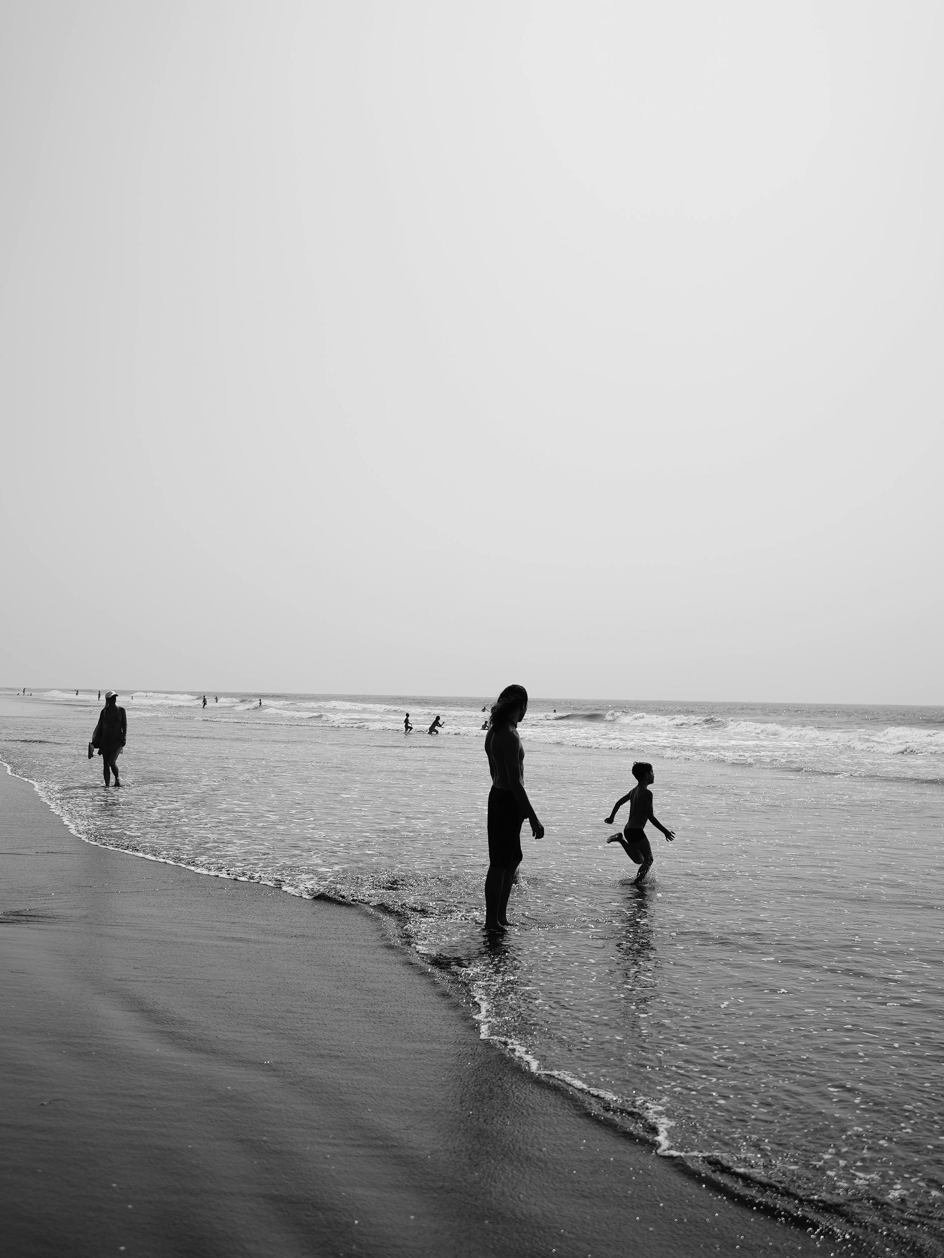 People wading and playing in shallow ocean water at a beach, with gentle waves and a cloudy sky.