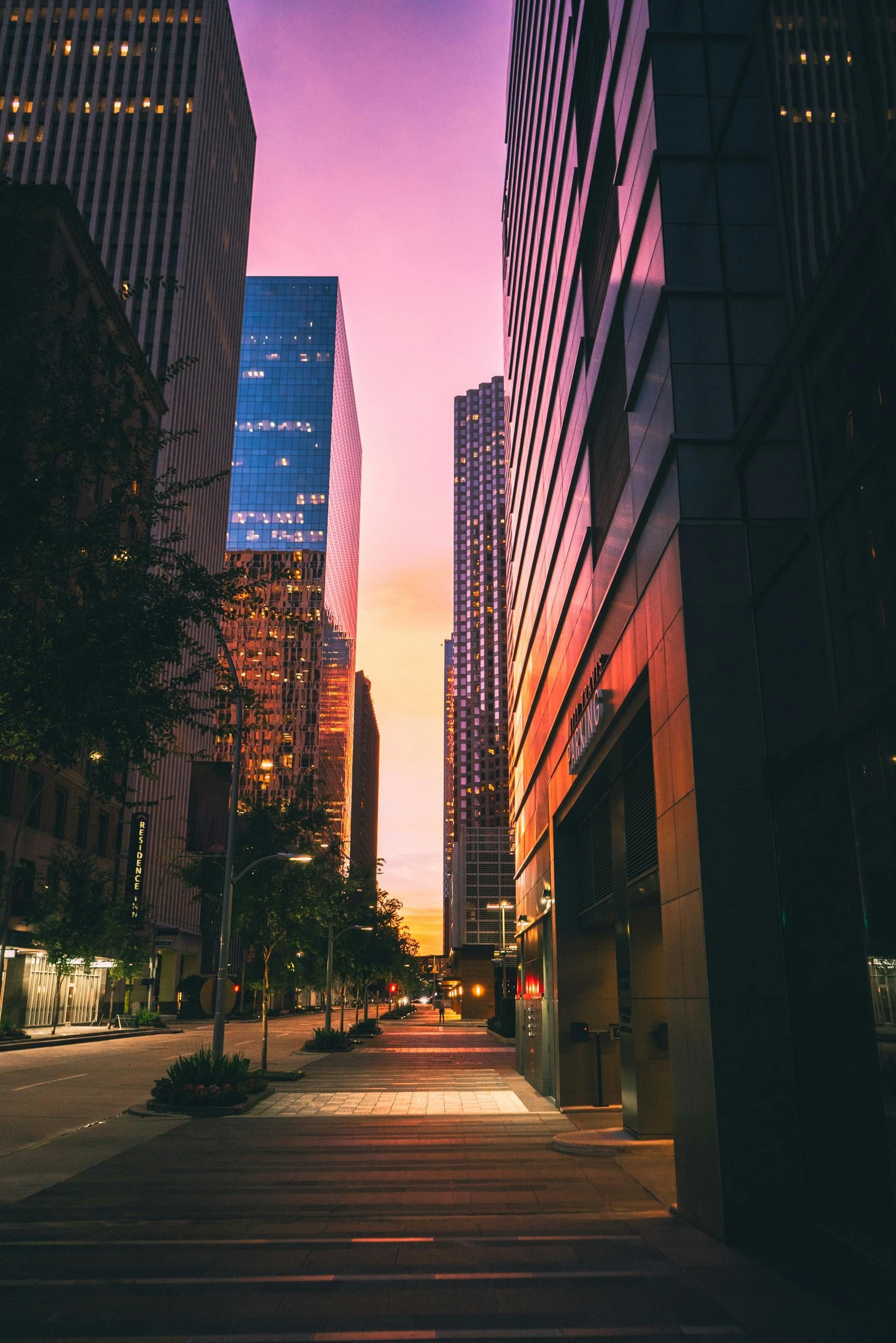 Sunset city street between tall glass buildings, with warm lights and a pink sky.