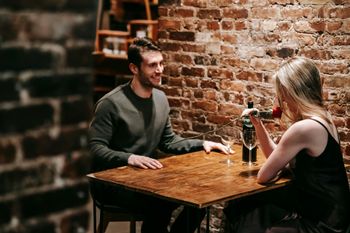 Two people talking at a small wooden table in a brick-walled cafe, with a beer bottle and flowers on the table