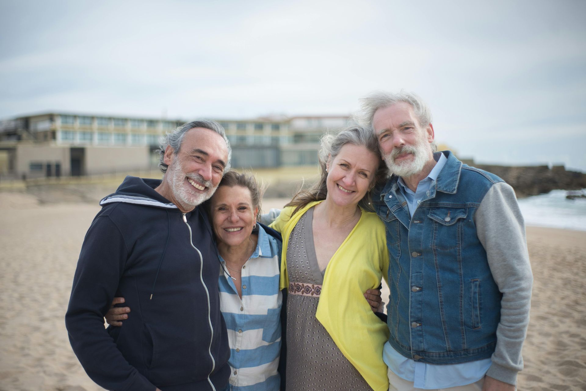 Four smiling people standing together on a beach, with buildings blurred in the background.