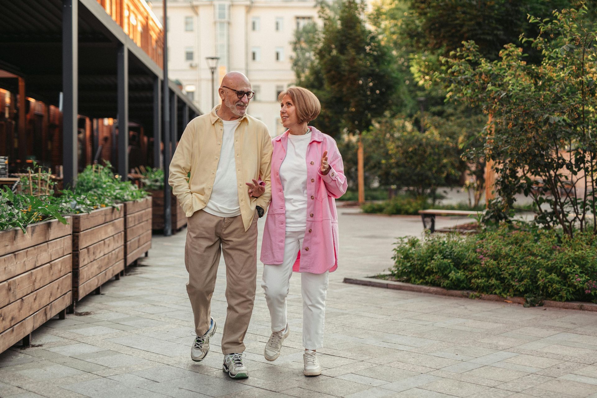 Two people walking and chatting on a tree-lined urban path beside outdoor cafés.