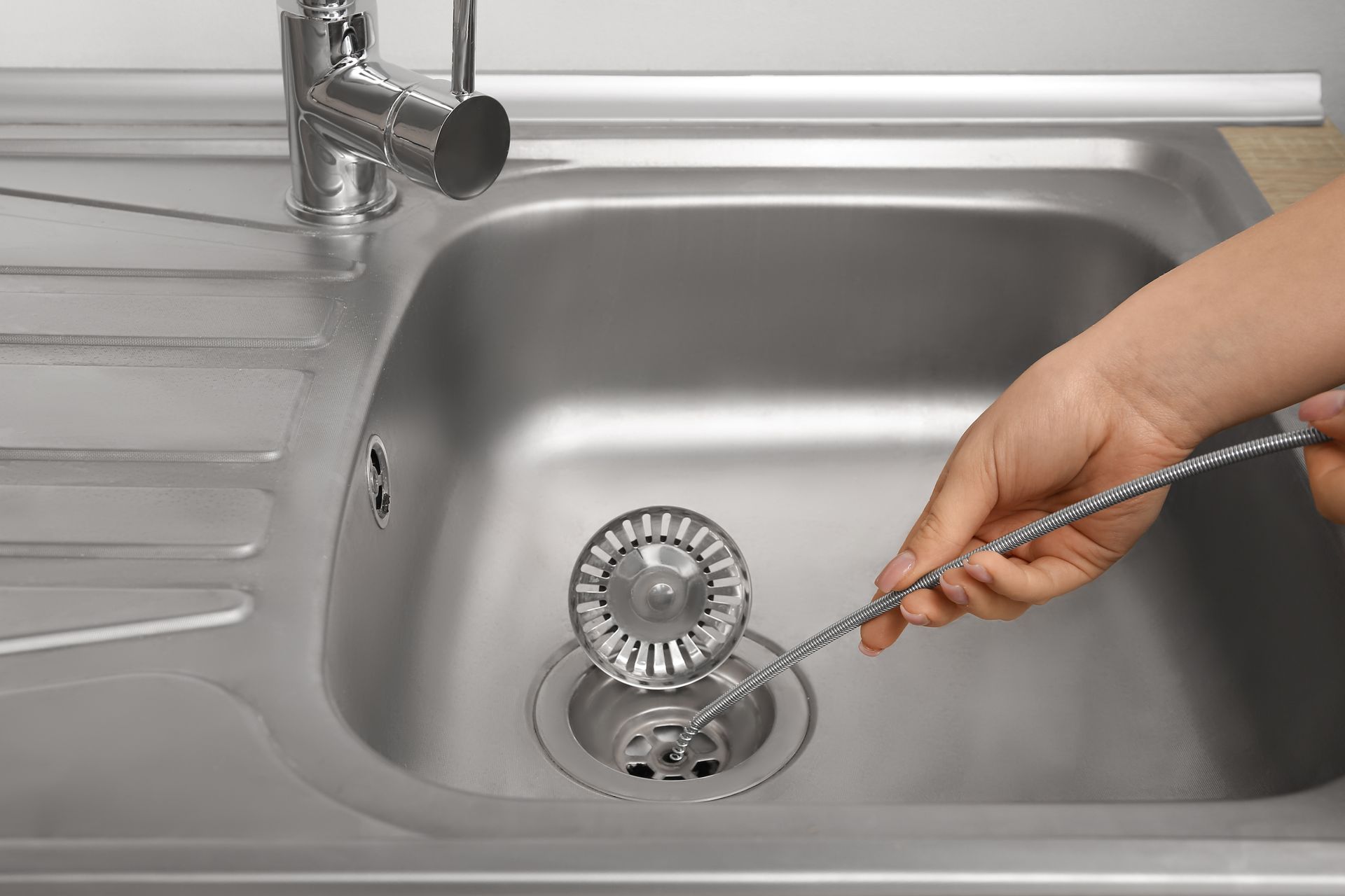 Hand using a drain snake to clear a stainless steel kitchen sink drain. Hand using a drain snake to clear a stainless steel kitchen sink drain.