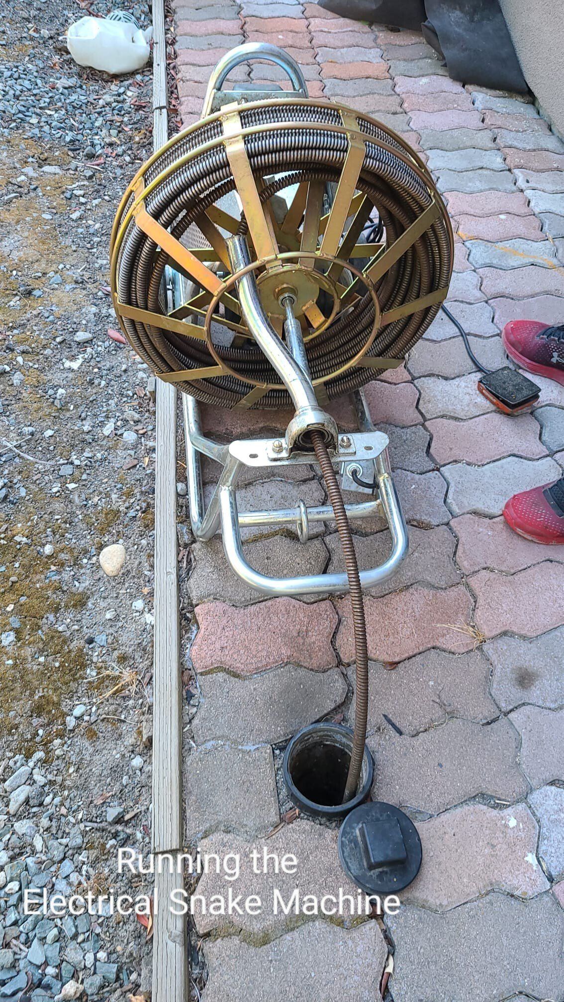 A gold, motorized drain cleaning machine on a paver patio with a metal snake cable feeding into an open sewer access pipe.