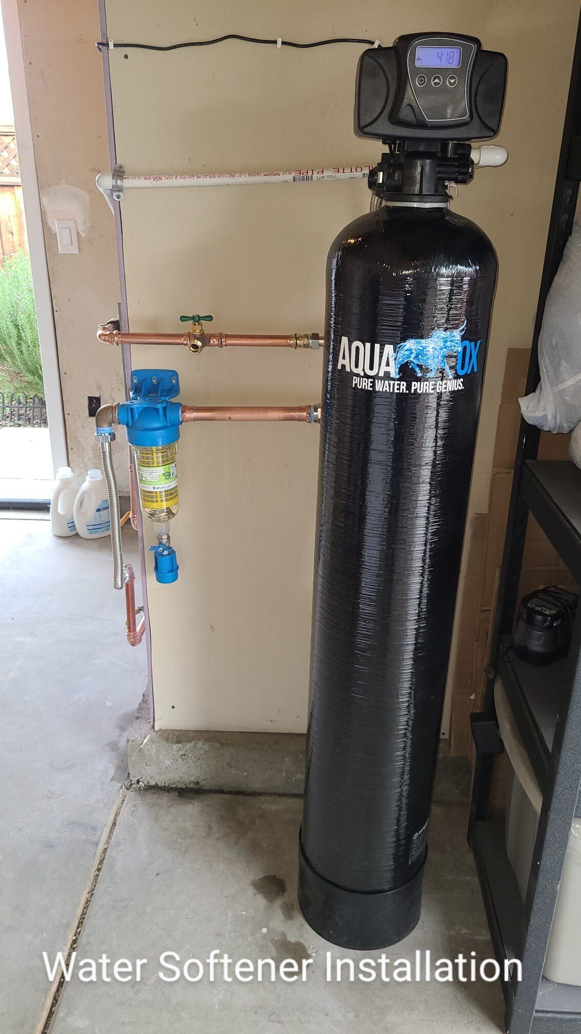 An AquaOx water softener system installed in a garage, featuring a black tank and a blue sediment filter on copper pipes.
