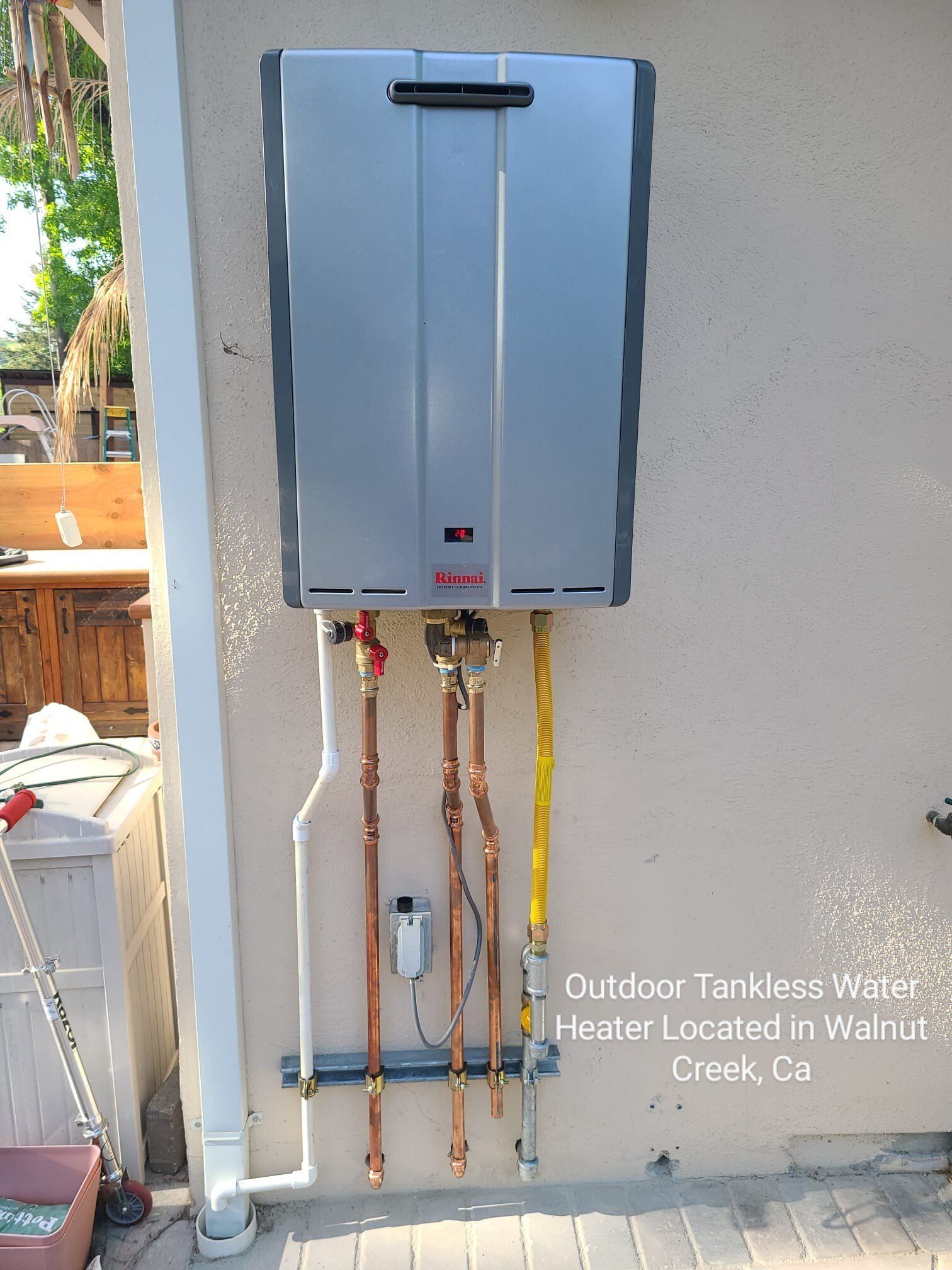 A wall-mounted, silver outdoor tankless water heater with visible copper water pipes and a yellow gas line.