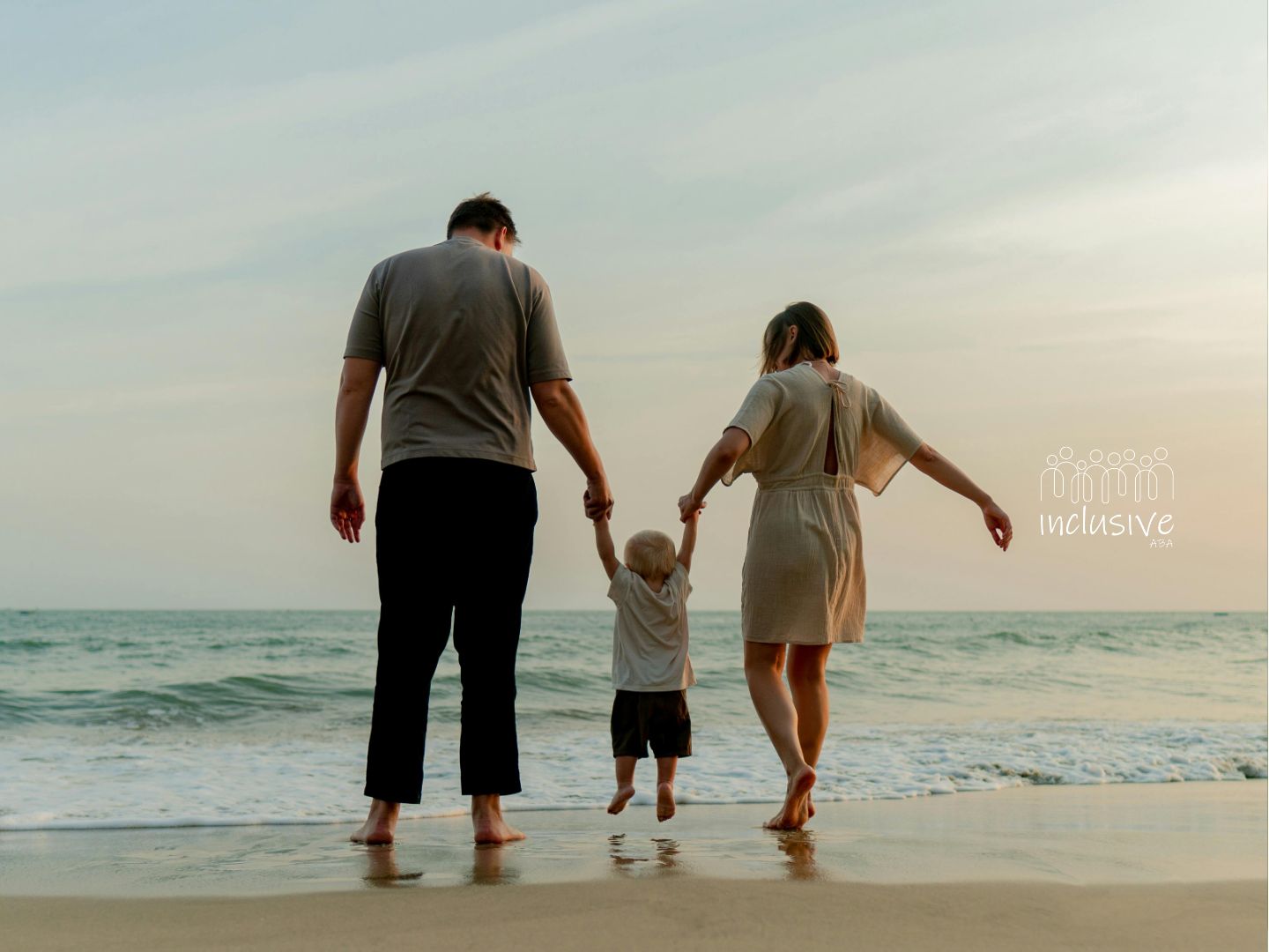 Family of three holding hands, walking along a beach at sunset.