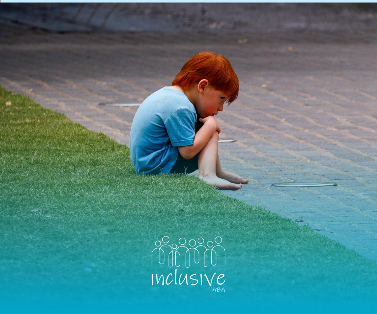 Young child in a blue shirt sits alone on the grass beside a paved path, knees pulled to chest.