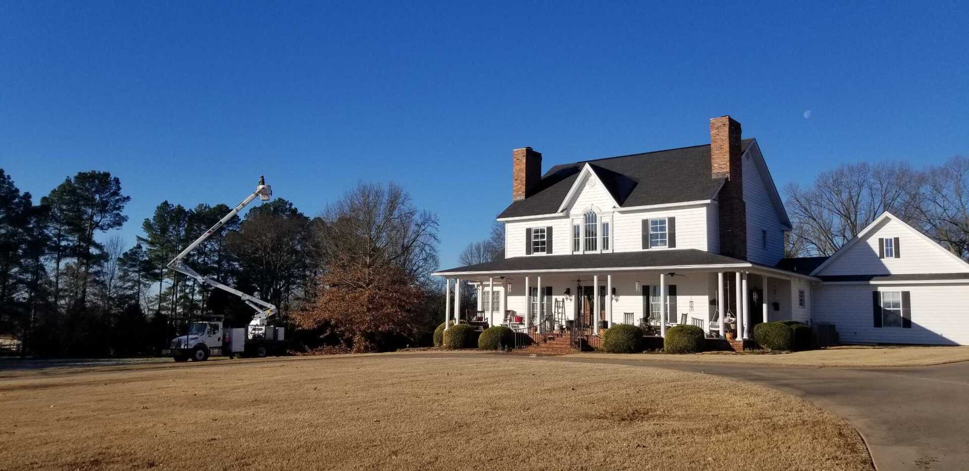 A white house with a wraparound porch and two chimneys, a tree service truck and clear blue sky.