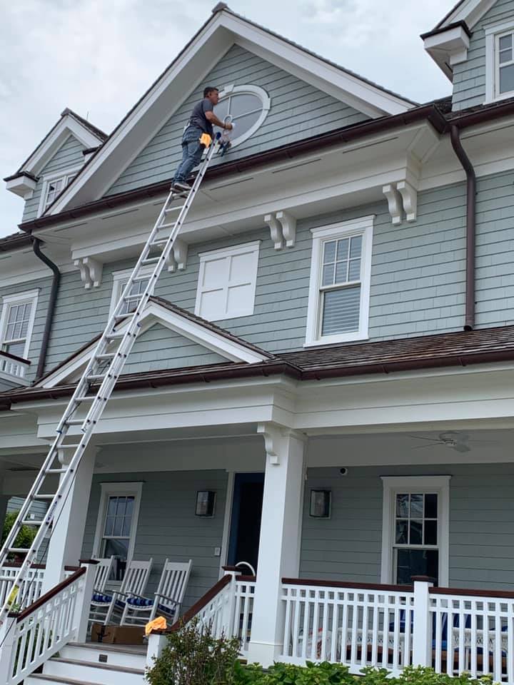 A person on an extension ladder cleaning a circular window on the second story of a light blue-sided house.